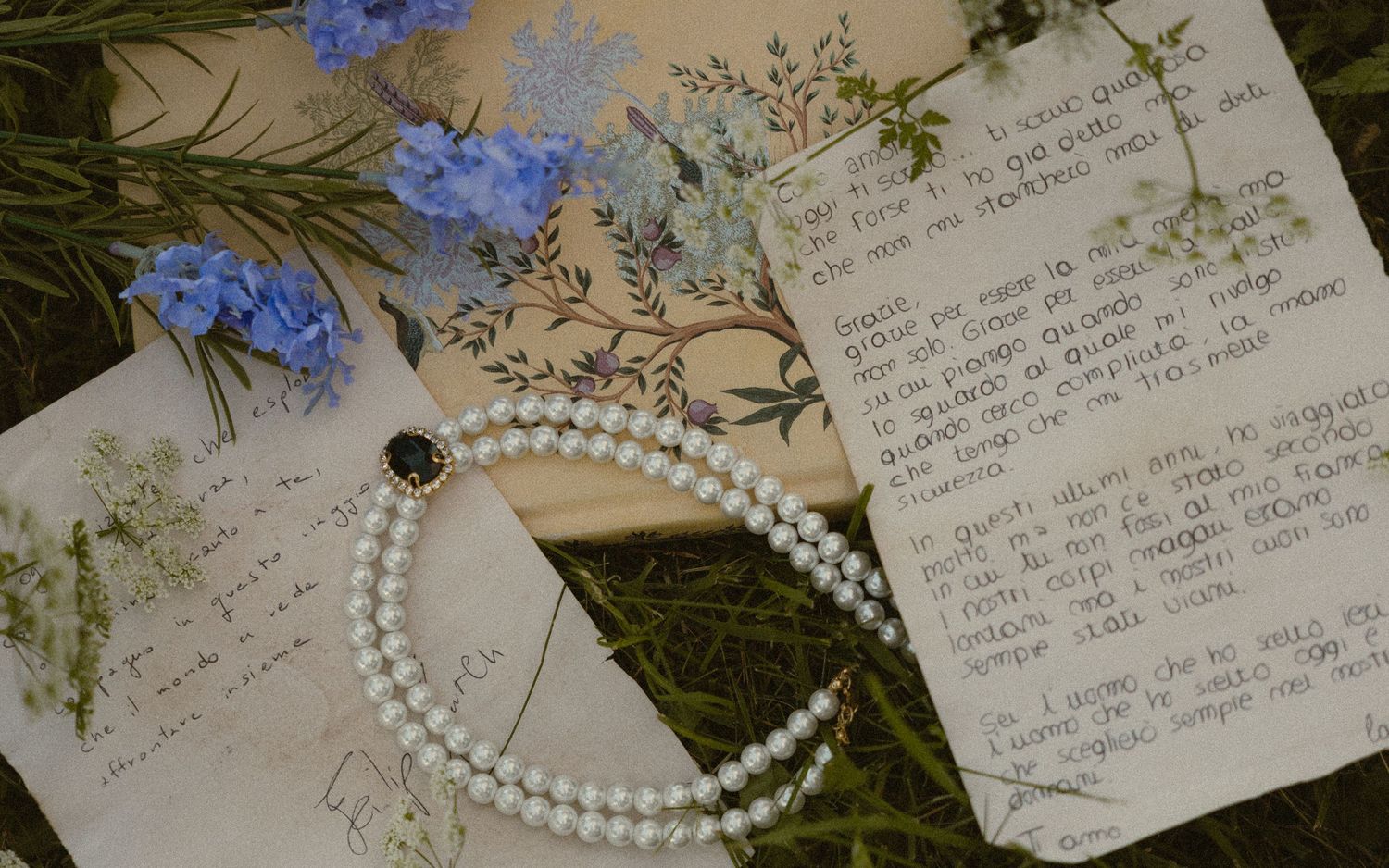 Vintage pearl necklace with dark centerpiece resting on handwritten letters and floral paper, surrounded by blue wildflowers and greenery.