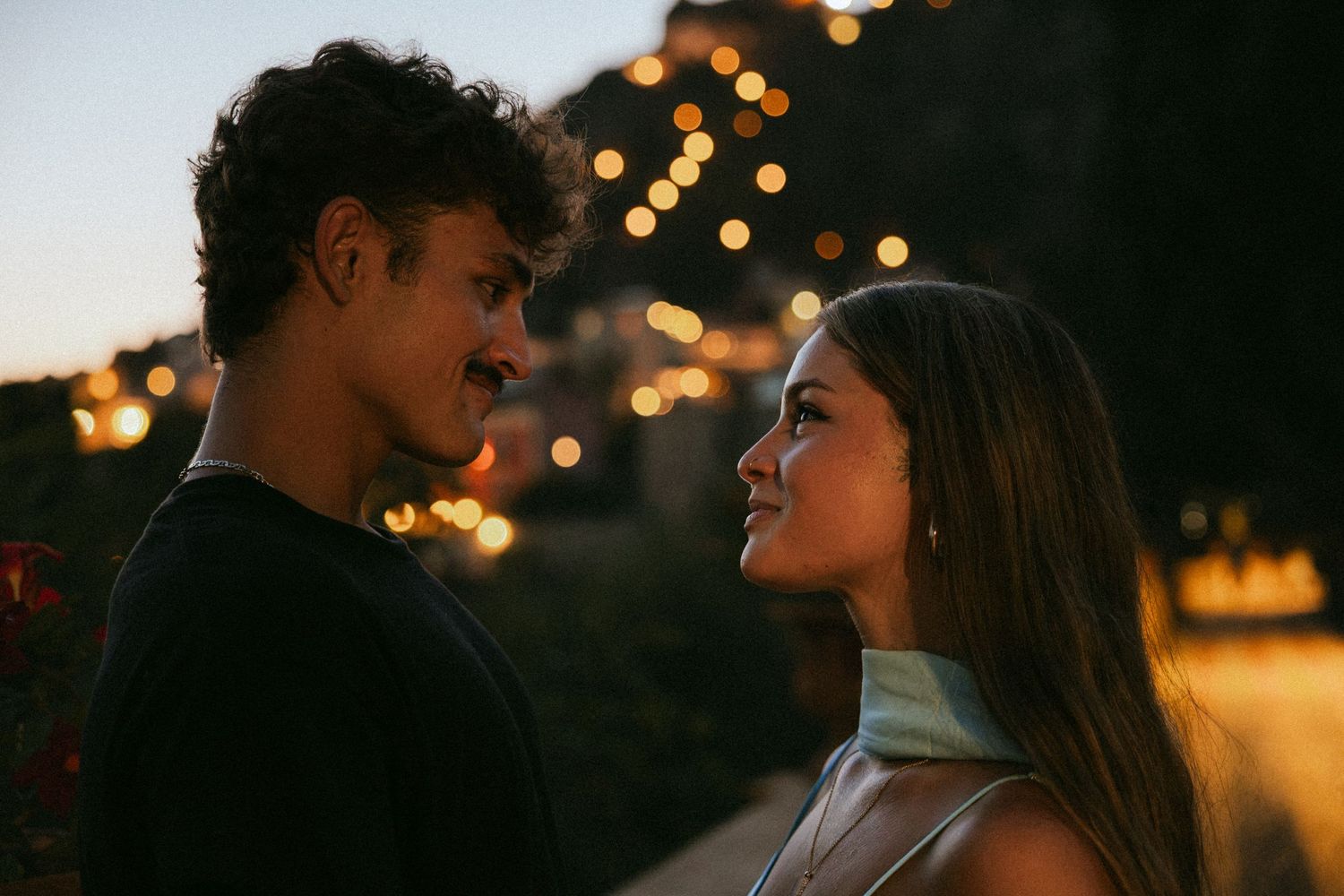 Young couple gazing at each other at dusk with warm glowing string lights and a hillside town twinkling in the background.