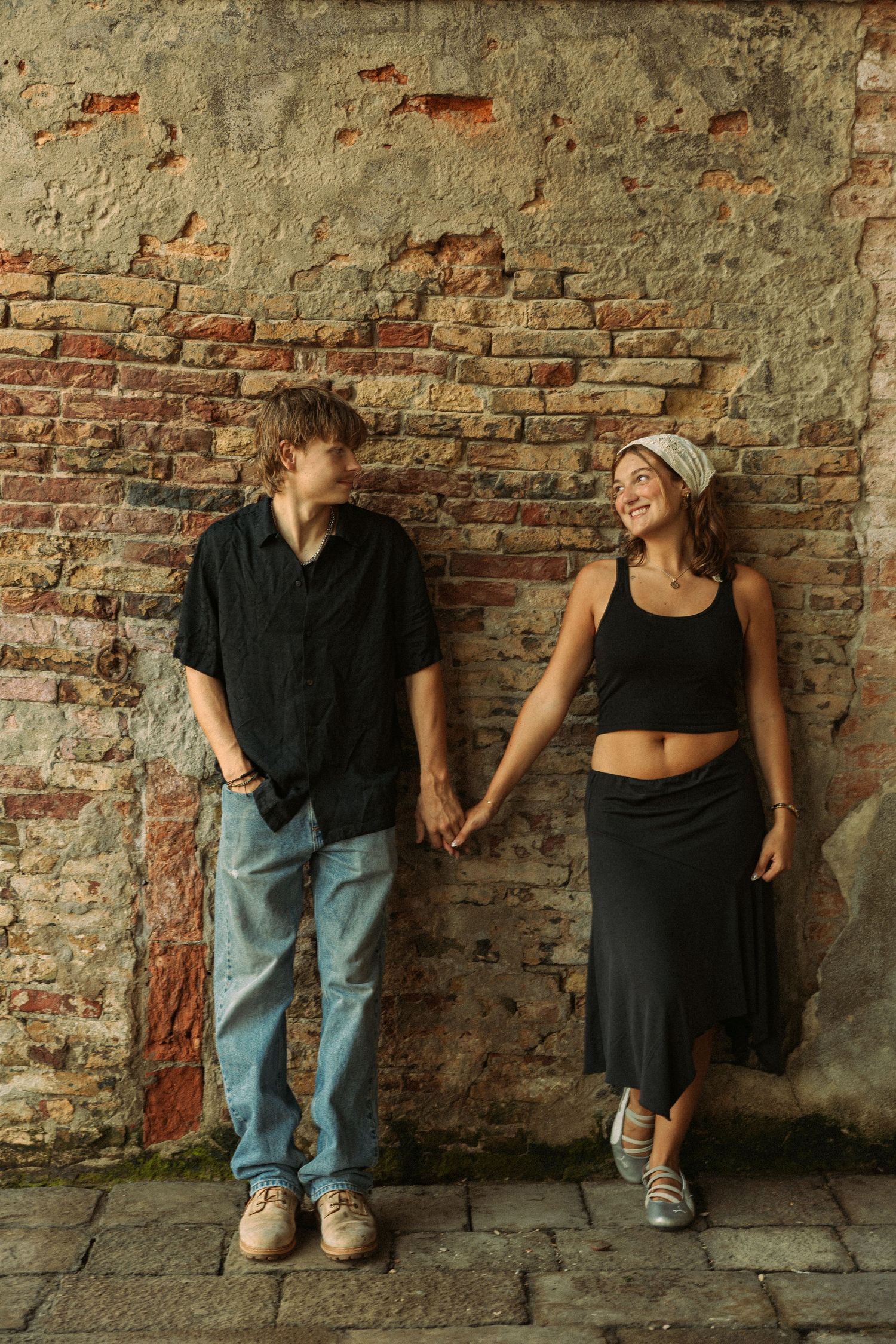 Young couple holding hands and smiling at each other while standing against a weathered, rustic brick wall on a cobblestone pavement.