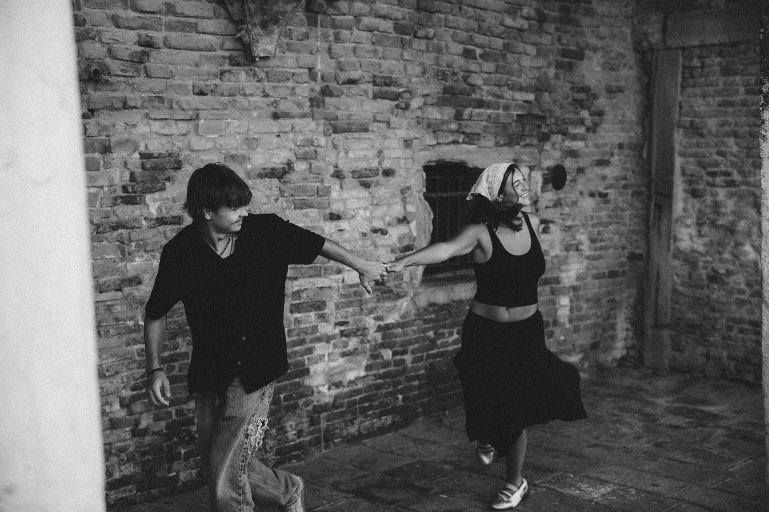 Black and white photo of a couple holding hands and dancing playfully in front of an old weathered brick wall.