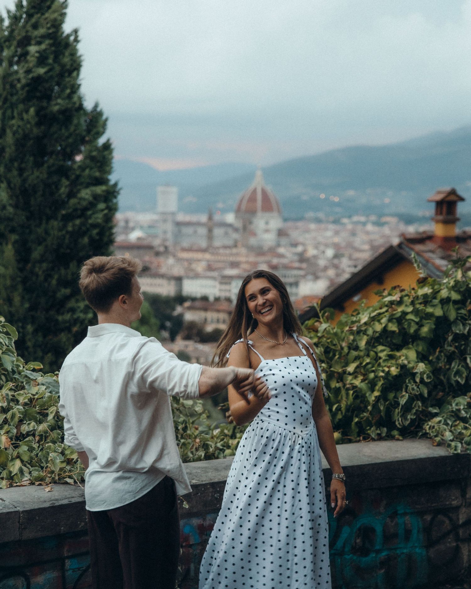 Smiling couple holding hands on a scenic overlook with a panoramic view of Florence, its red cathedral dome and misty hills behind.