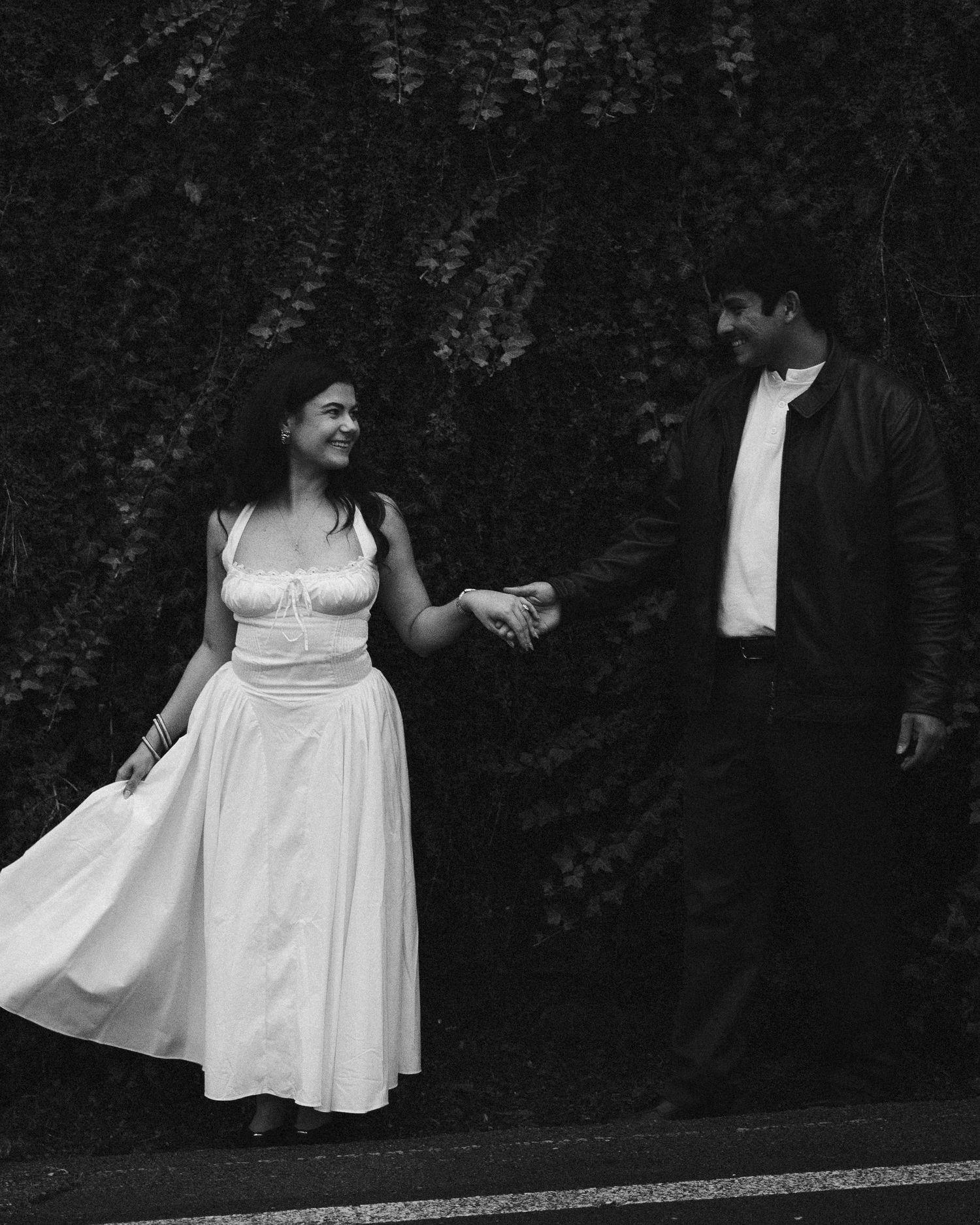 Black and white image of a couple holding hands and smiling at each other, the woman's white dress flowing, on a quiet road. ROMANTIC ITALY