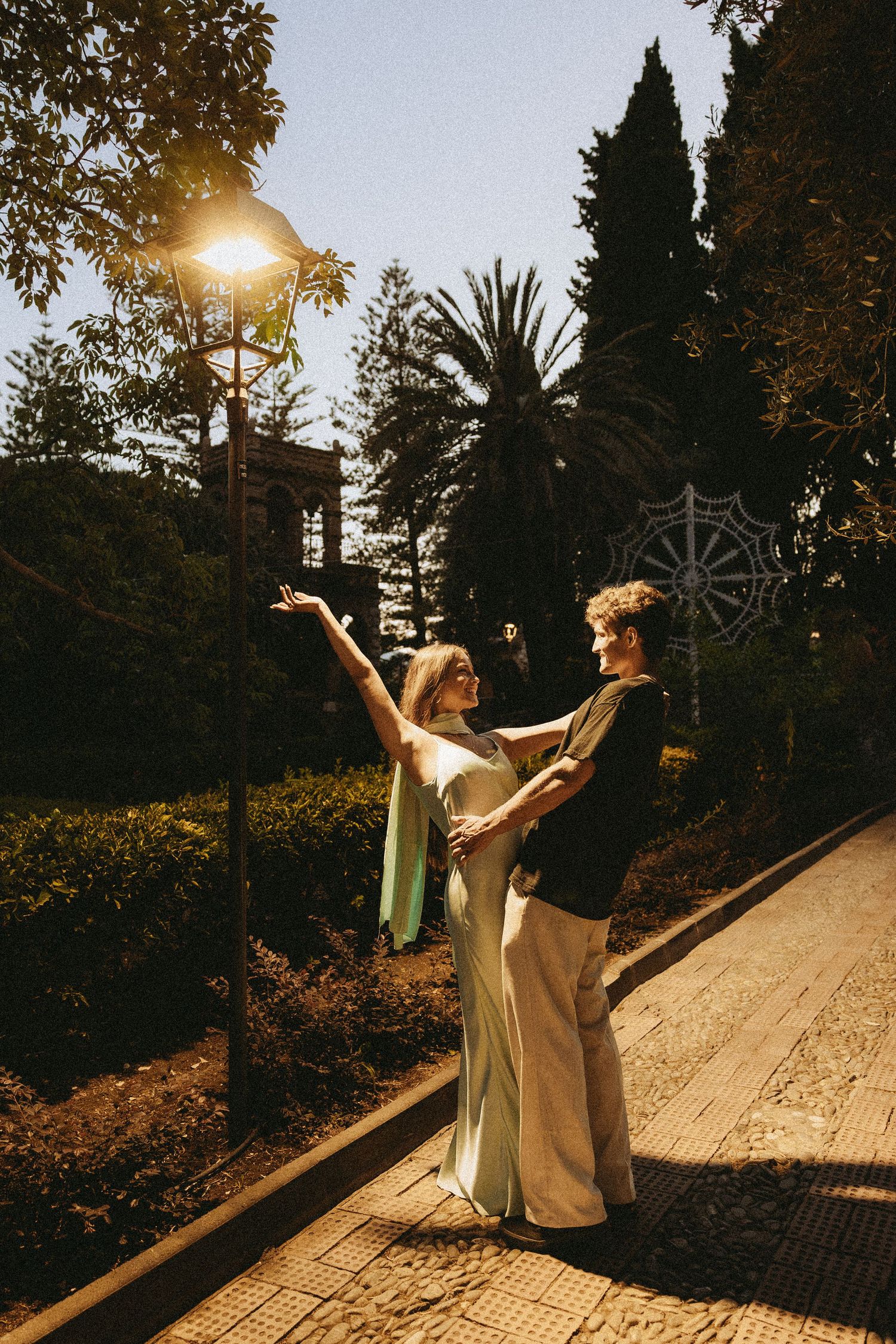 Two people in elegant evening wear dance on a stone path beneath a glowing street lamp, surrounded by palm trees and lush greenery at dusk.