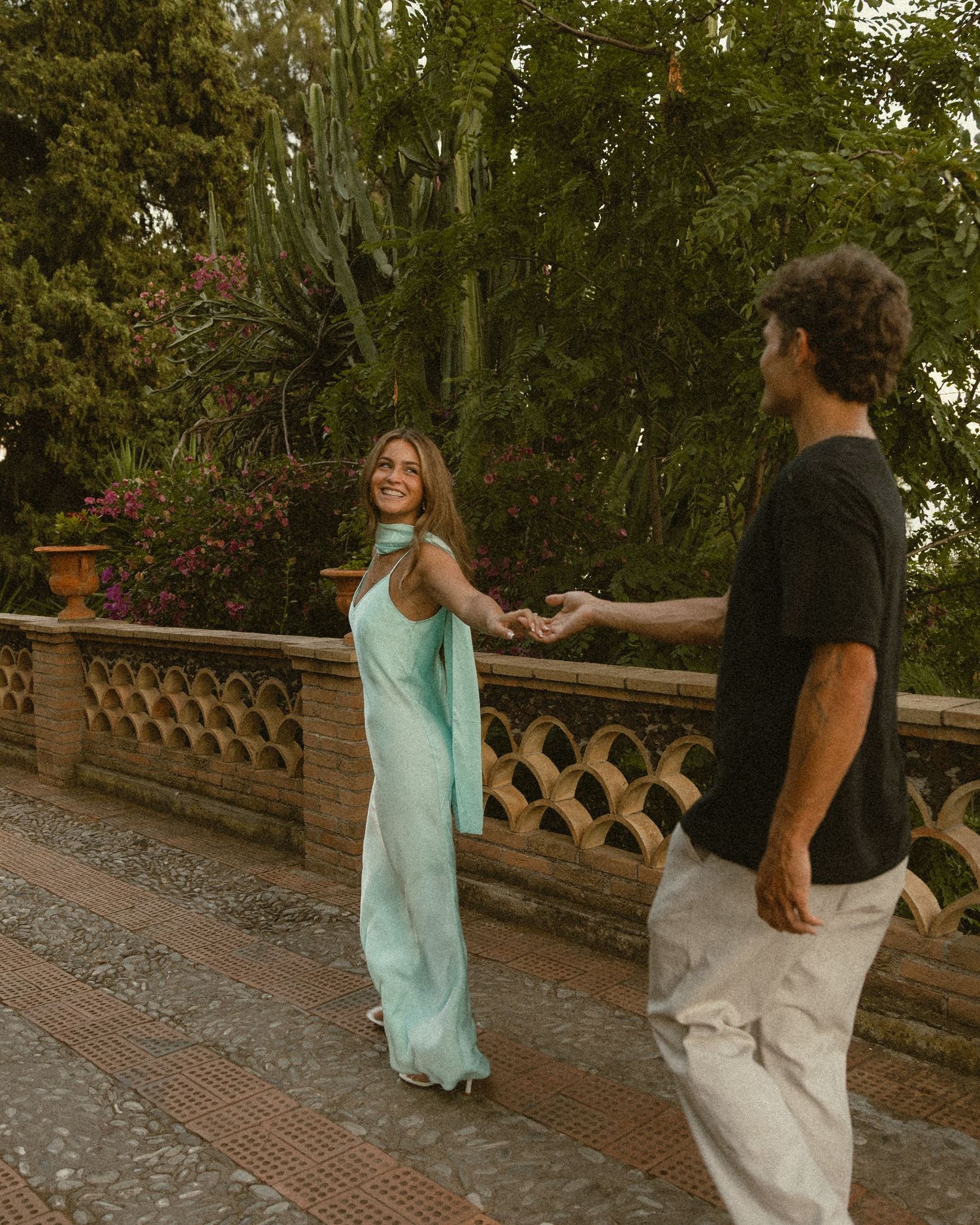 Woman in a flowing mint green dress and scarf holds hands with a man in a black tee while walking on a garden terrace.