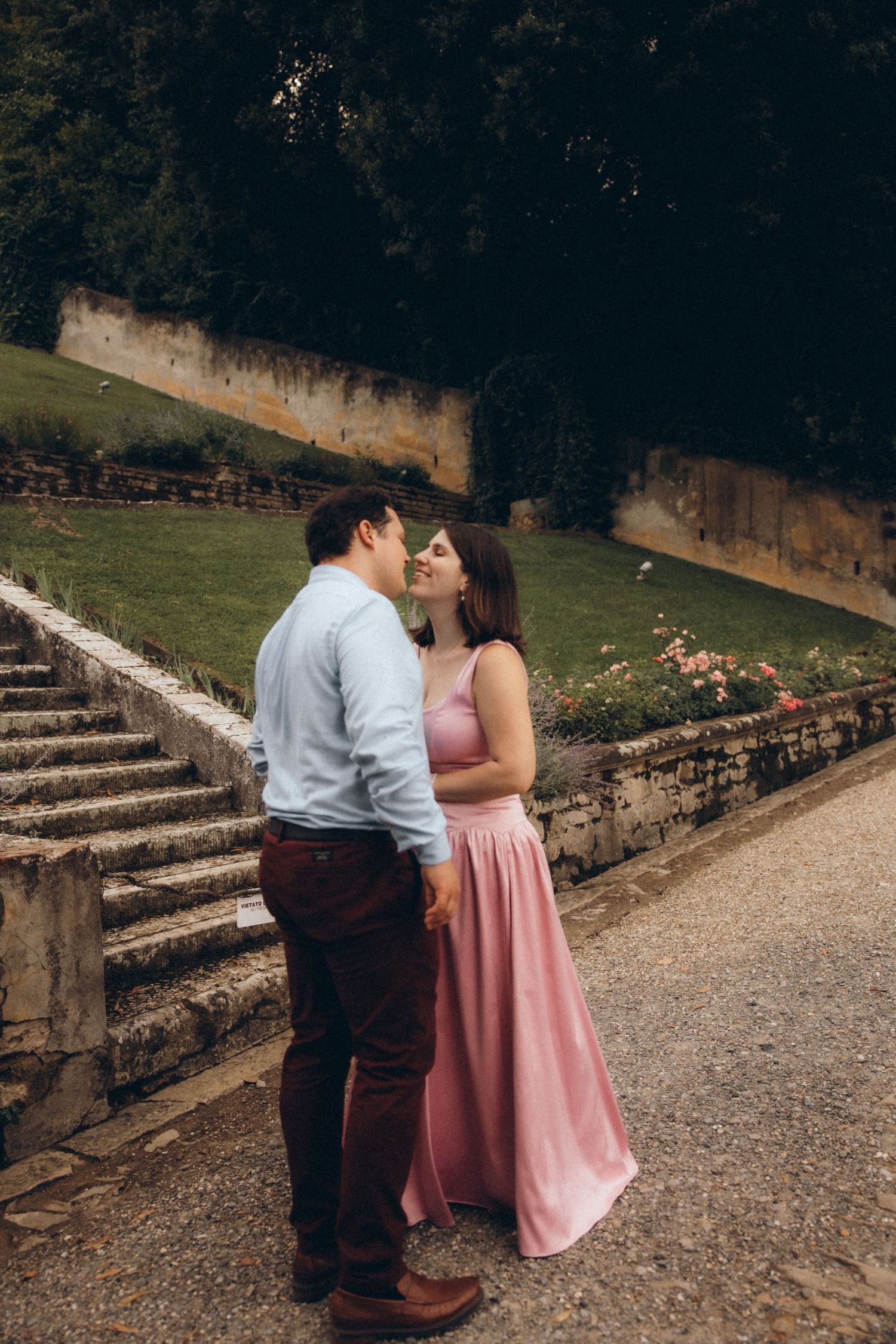 Couple in a pink satin gown and light blue shirt share an intimate moment on a gravel path beside stone steps and a lush garden.