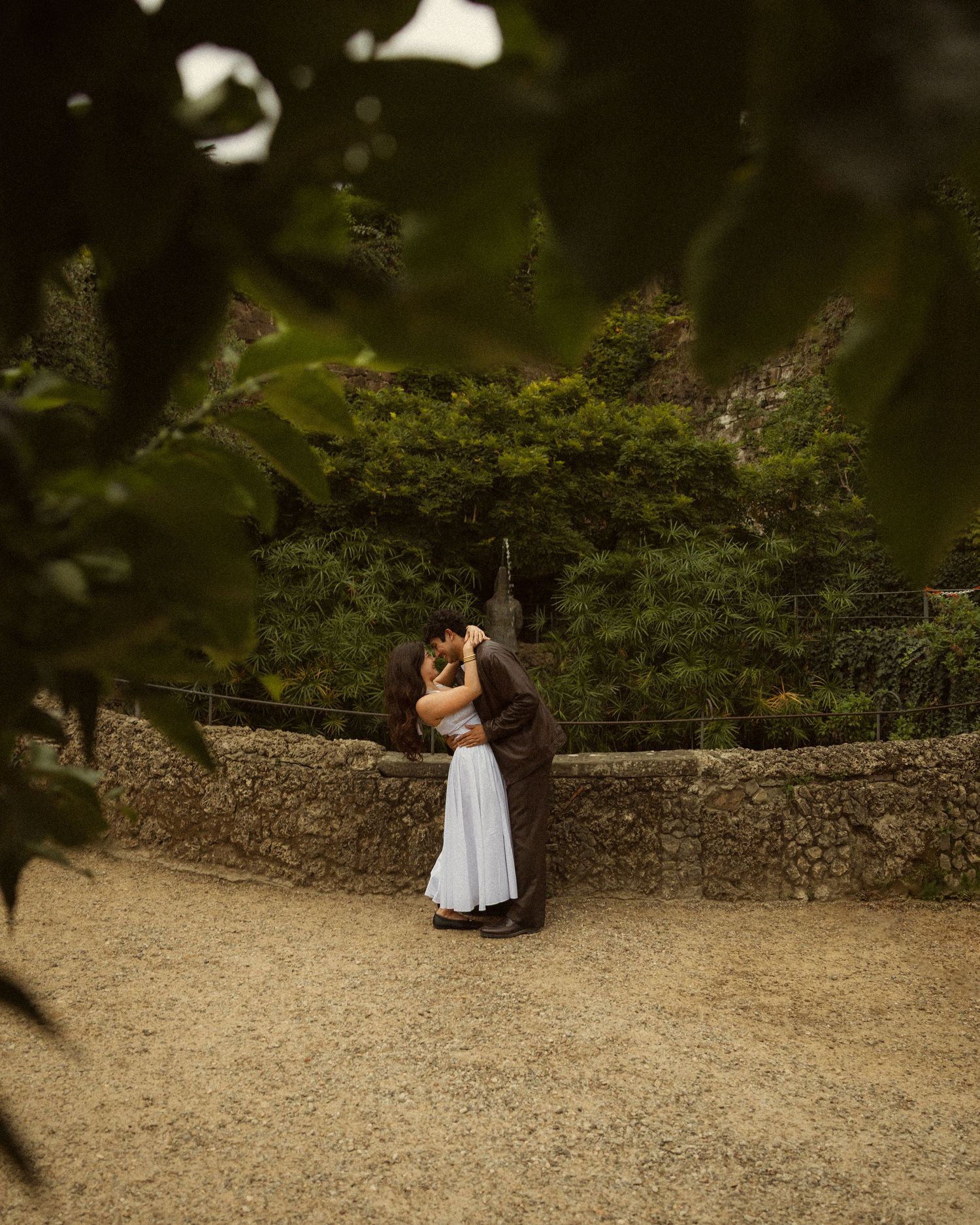 Couple in white dress and dark suit embracing and kissing beside a stone wall, framed by green leaves in a lush garden setting.