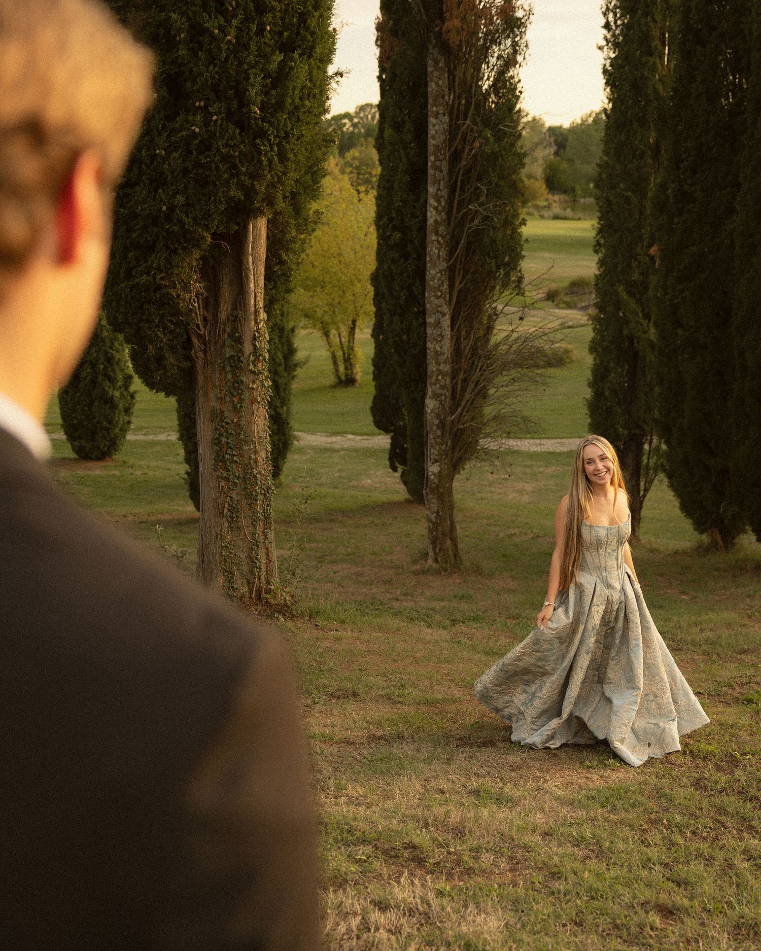 Woman in a flowing sage green gown stands on a grassy lawn framed by tall cypress trees as a man in a dark suit watches from the foreground.