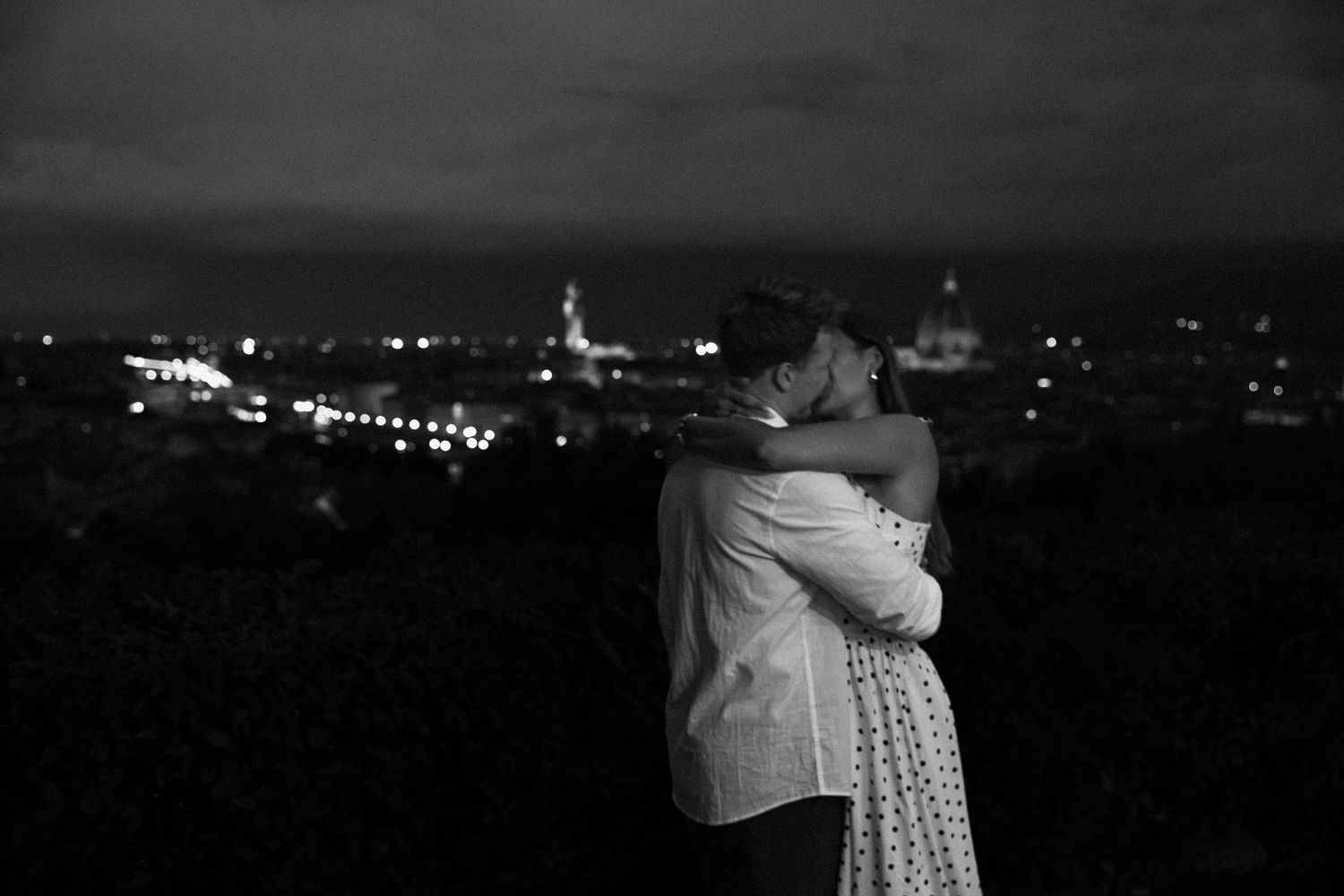 Black and white photo of a couple embracing and kissing on a hillside overlooking a glowing city skyline and domed landmarks at night.