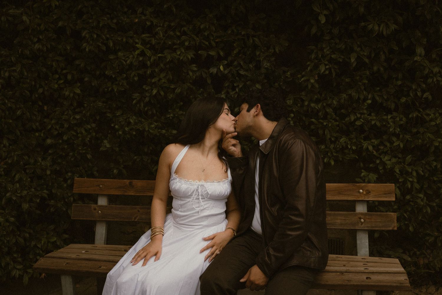 Couple sharing a romantic kiss on a wooden park bench, the woman in a white dress and the man in a dark jacket, before a dark green hedge.