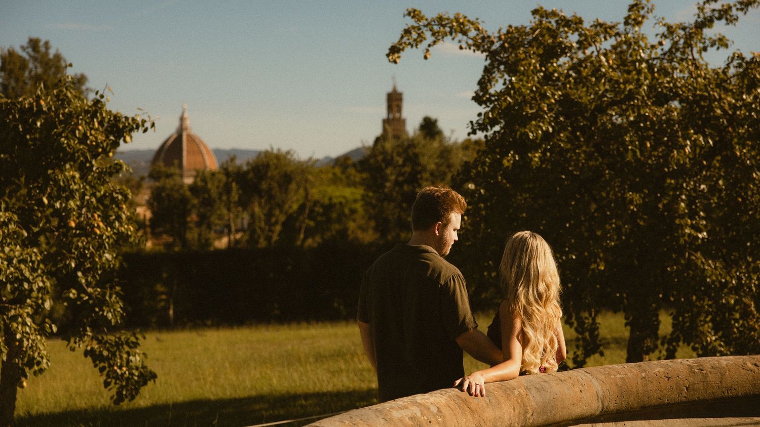 Couple standing close at a stone wall overlooking a sunlit Florence garden with the cathedral dome and tower in the distance.