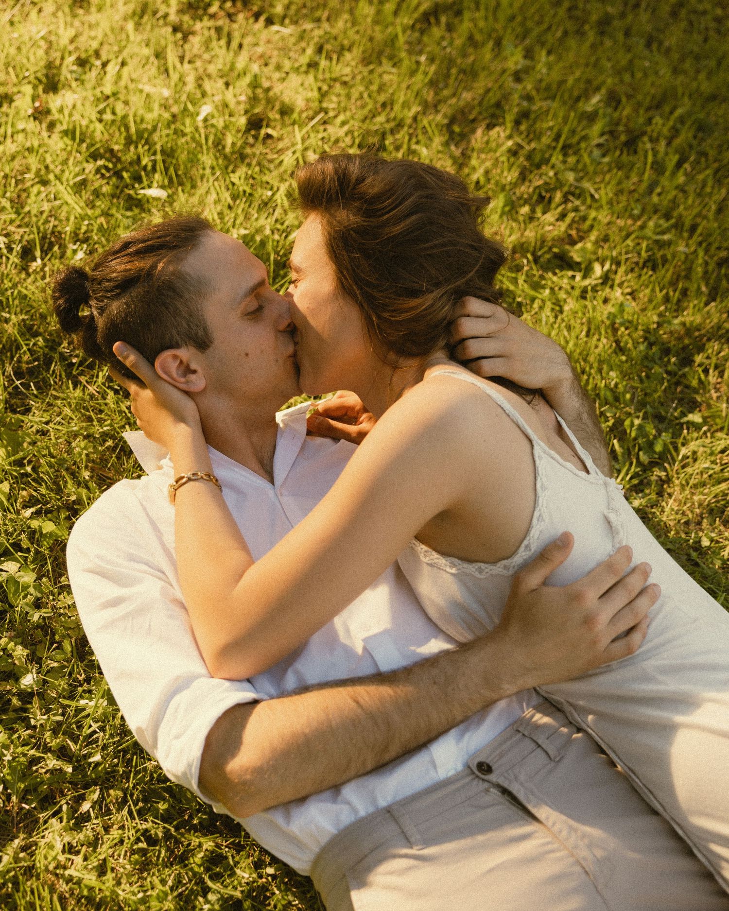 A couple in light clothing lies on sunlit green grass, embracing and kissing in a romantic outdoor moment.