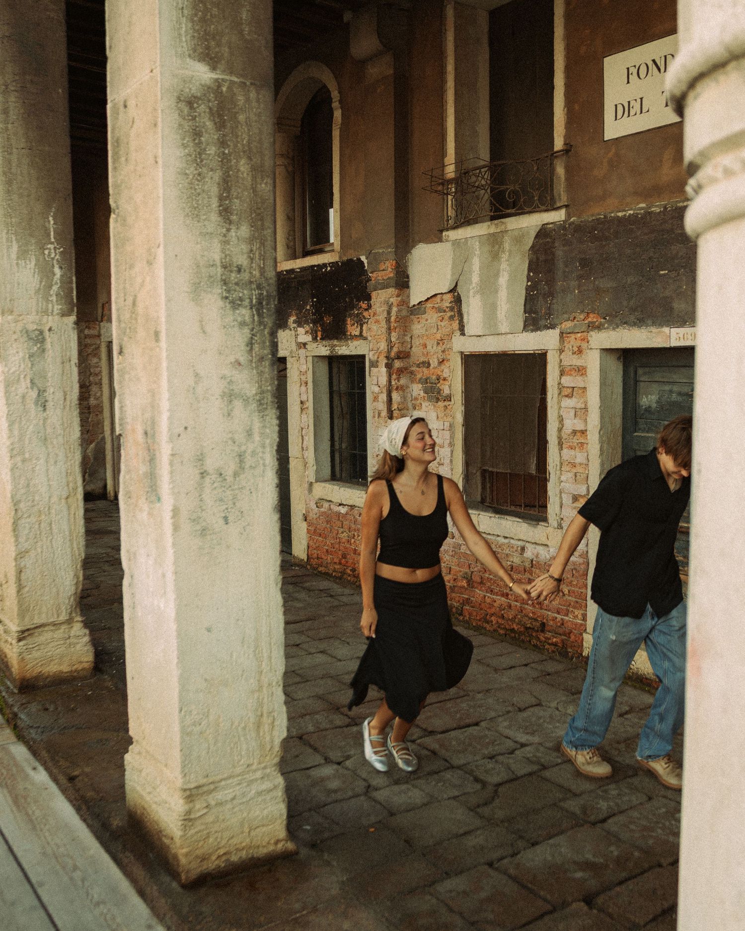 Smiling woman in a black top and skirt walks hand in hand with a man along a historic Venetian arcade with stone columns.