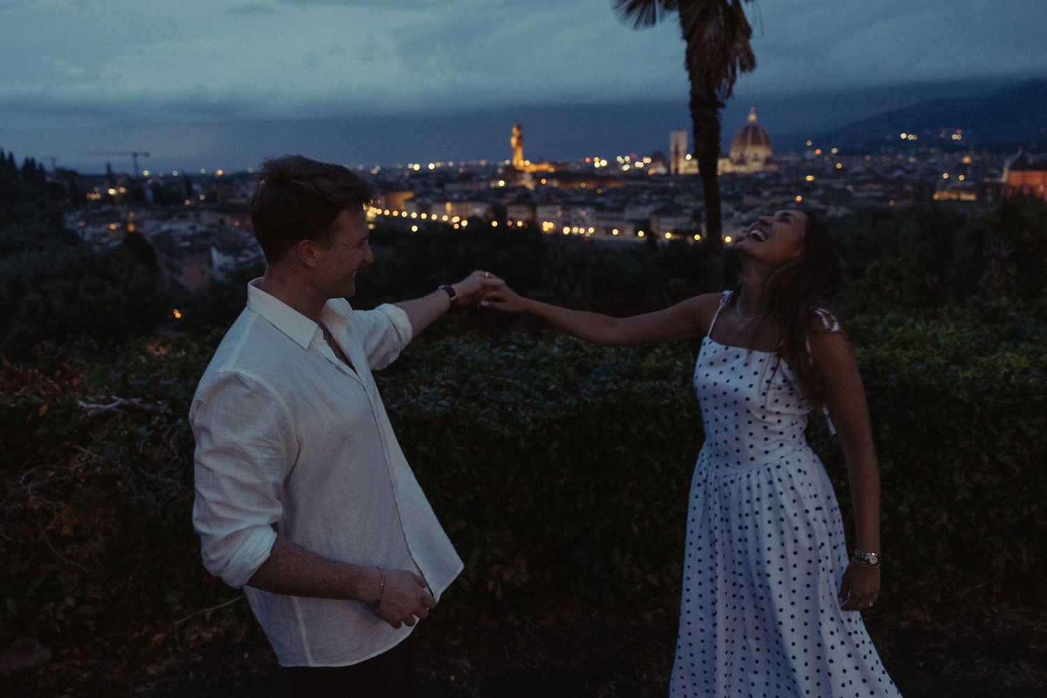 Couple holding hands and dancing at dusk on a hillside overlooking a glowing city skyline, with a palm tree and twilight sky behind them.