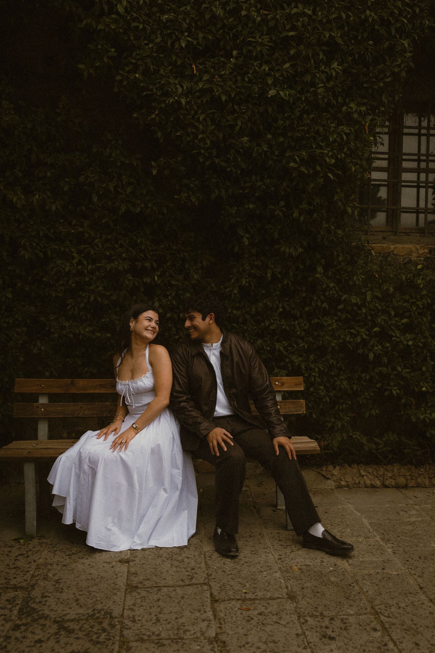 Smiling couple sits close on a wooden bench in front of a green ivy-covered wall, she in a white dress and he in a brown jacket.