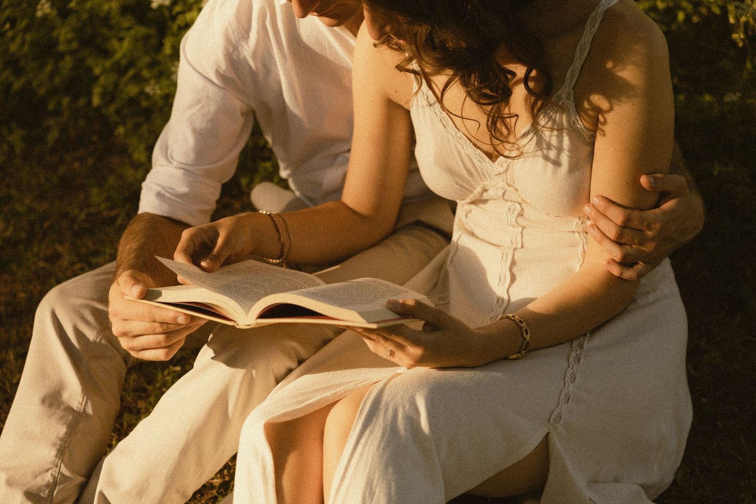Couple in white linen outfits sit close together outdoors in warm golden light, reading an open book on their laps.