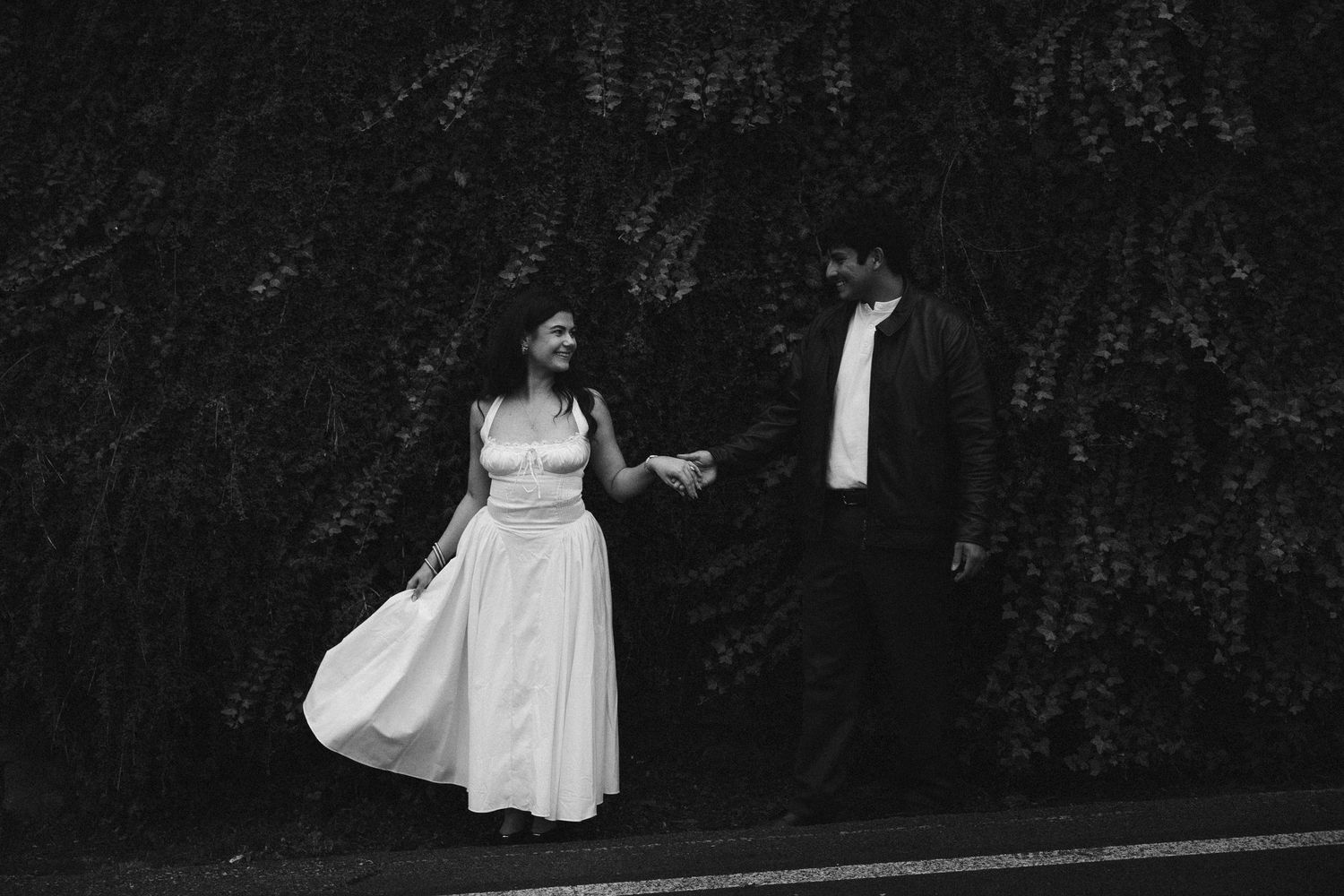 Black and white photo of a couple holding hands and walking along a road in front of a dark leafy wall, the woman in a flowing white dress.