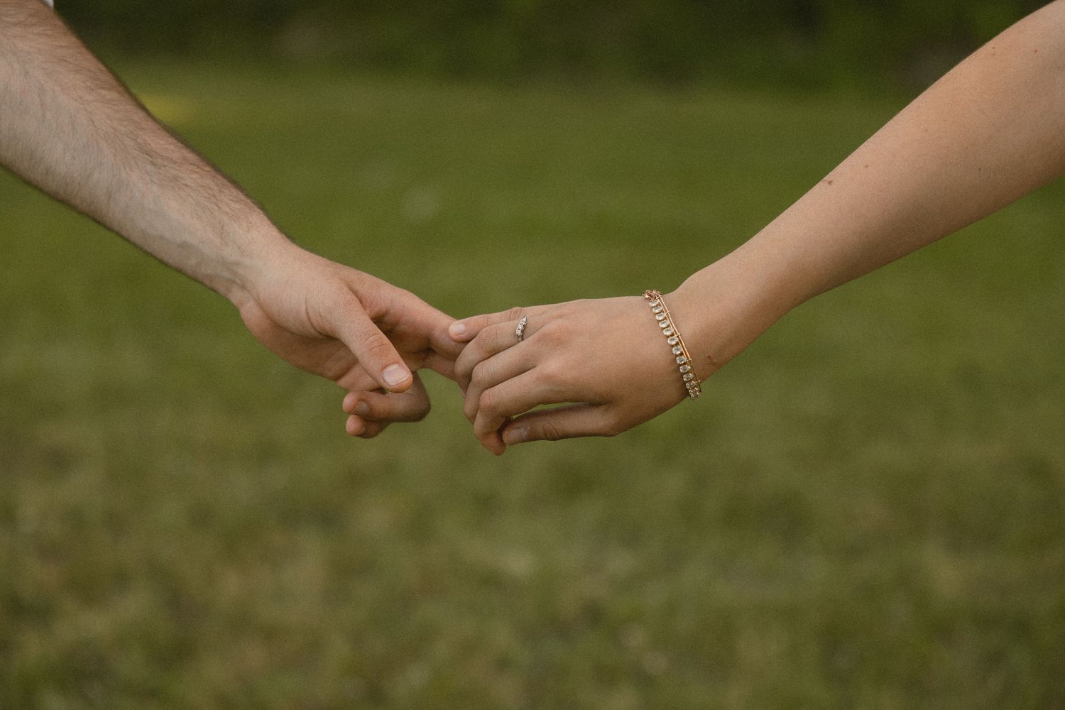 Two people gently holding hands outdoors on a grassy field, one wearing a gold bracelet and a ring.
