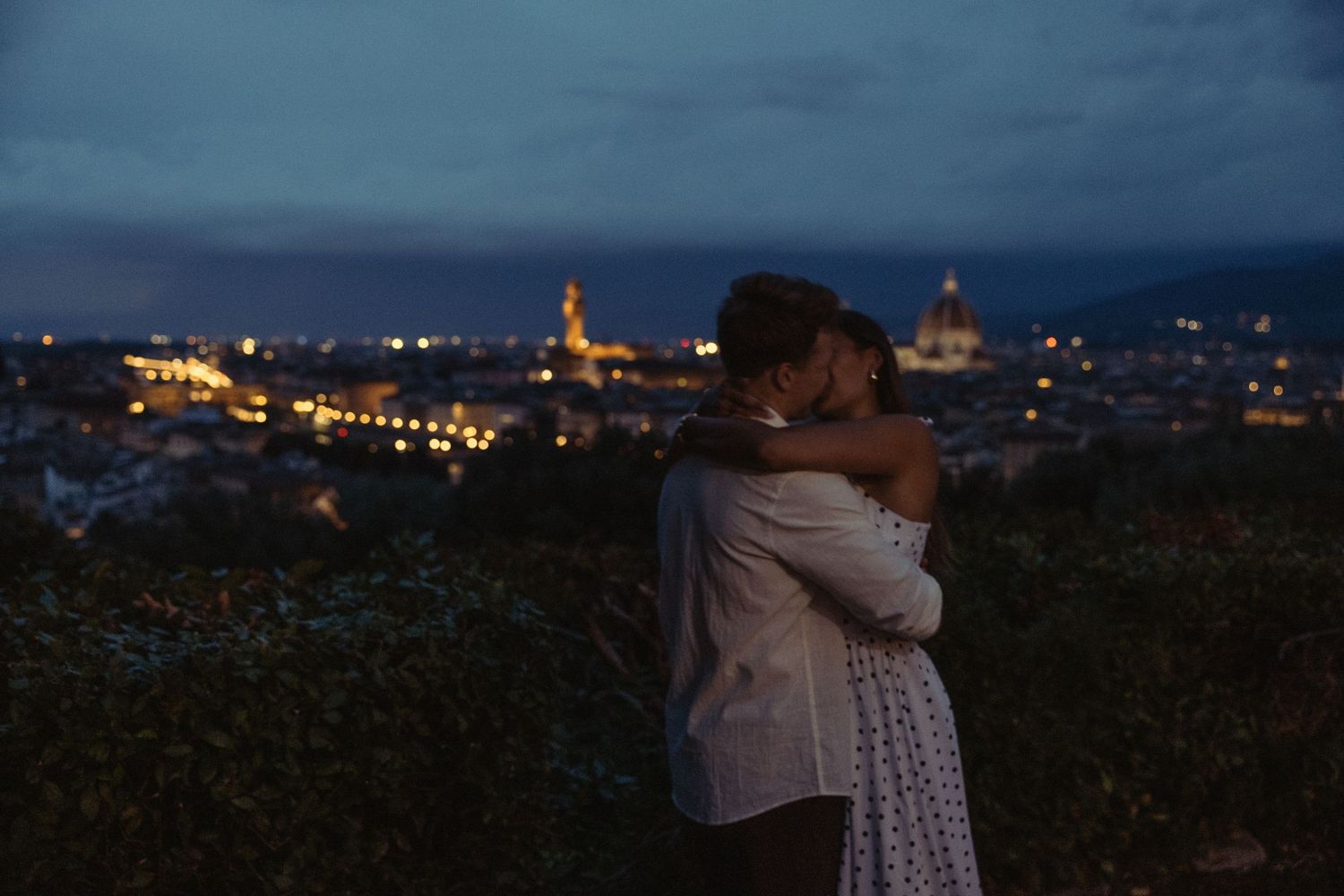 Couple embracing at dusk on a hilltop overlooking the glowing city lights and skyline of Florence, Italy.
