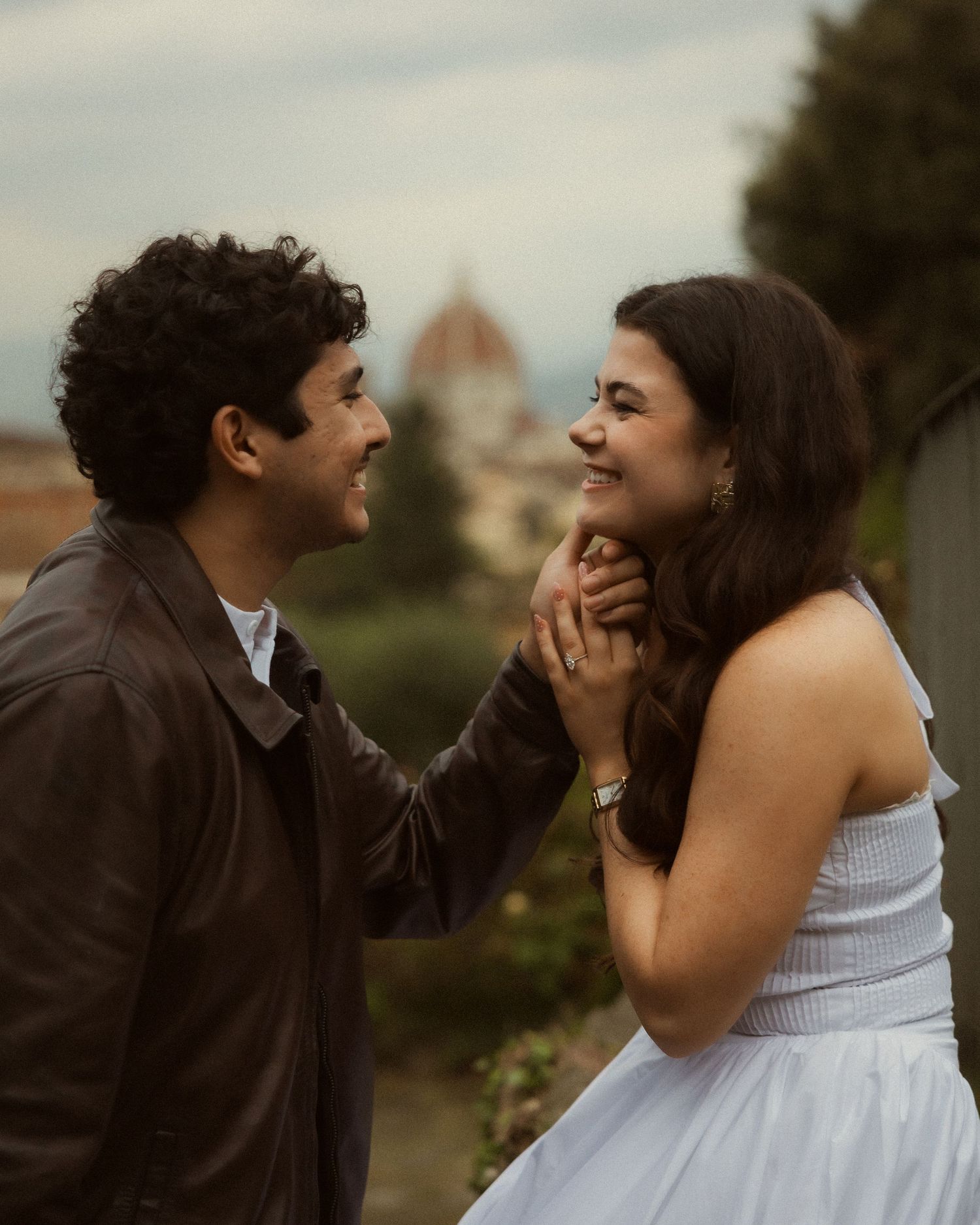 Smiling couple share a tender moment outdoors, the woman in a white dress holding the man's hand with a domed Florence skyline behind them.