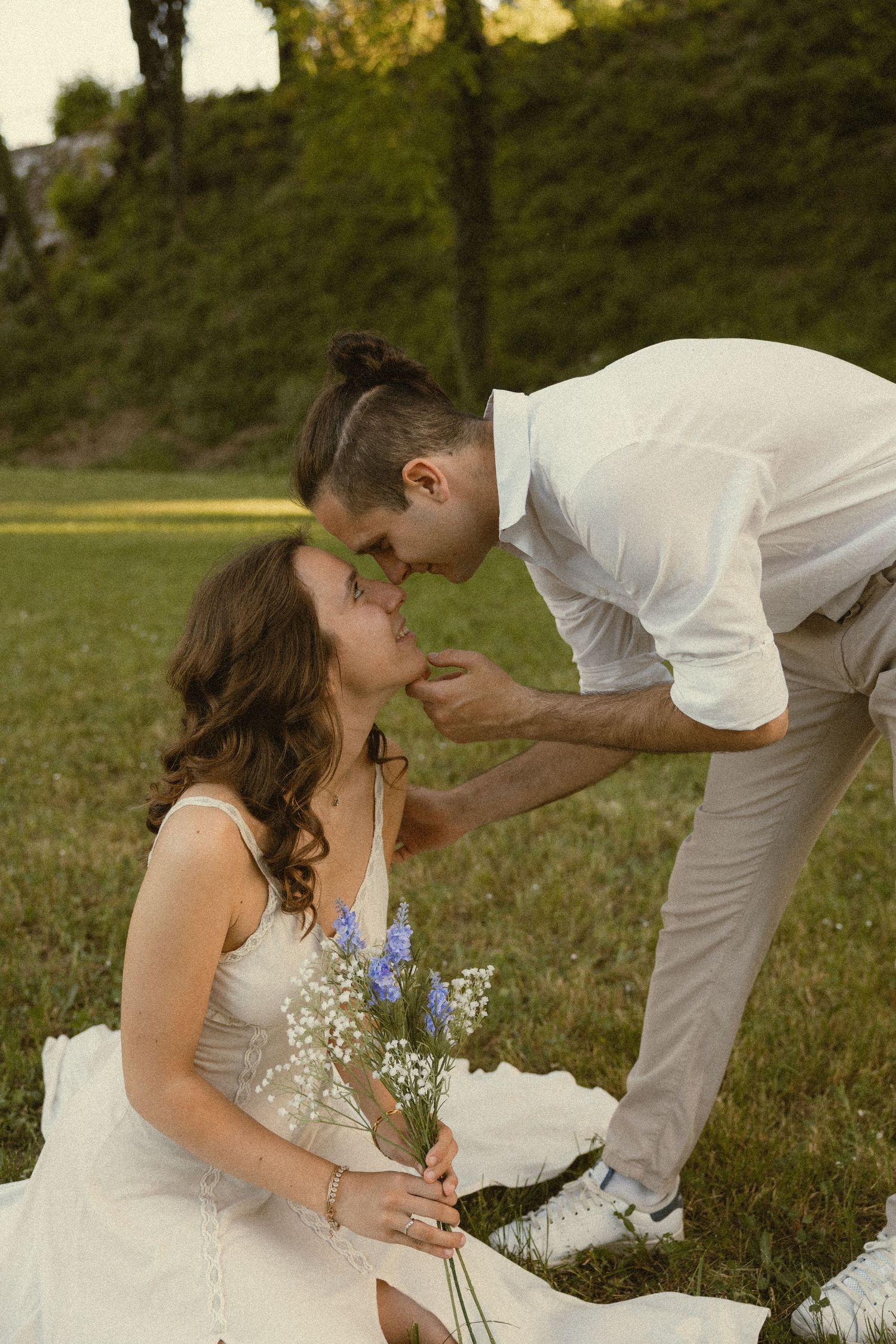 A man in a white shirt leans down to touch noses with a seated woman holding wildflowers on a sunlit grassy hillside.