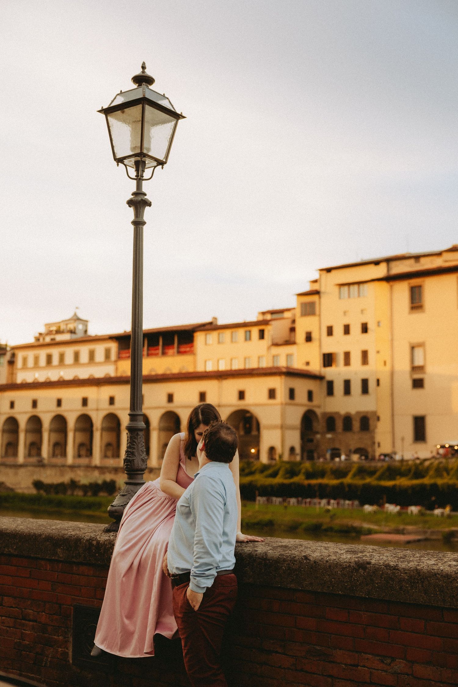 Couple embracing on a brick riverside wall beside a vintage street lamp with golden Florence-style buildings glowing behind them at sunset.