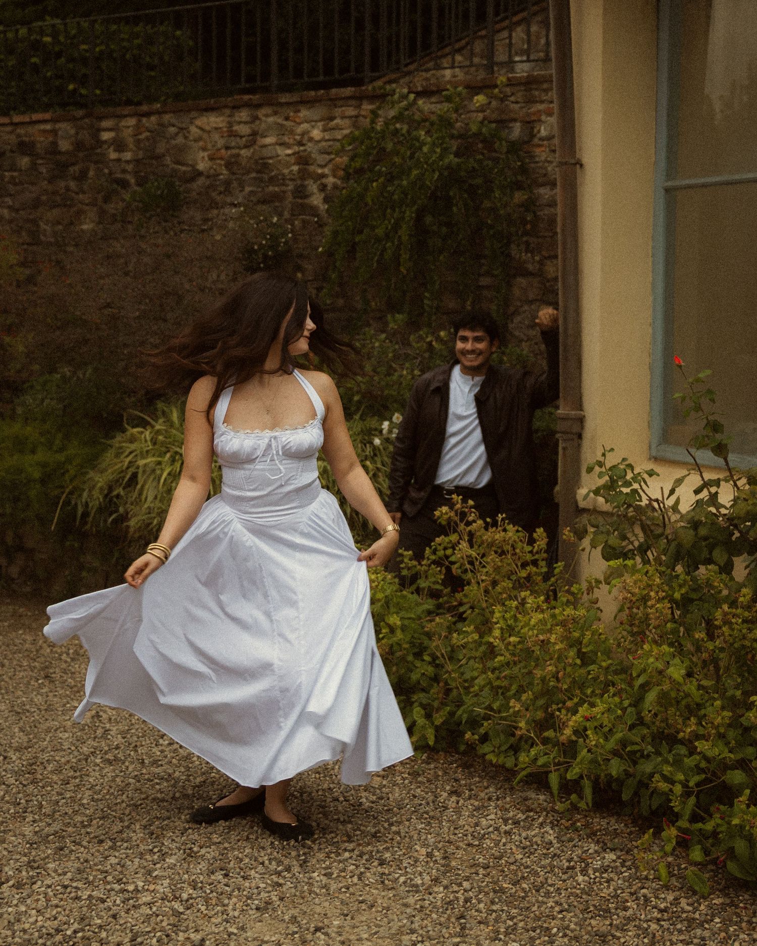 Woman in a flowing white halter dress twirls on a gravel path in a garden while a smiling man in a dark jacket dances behind her.