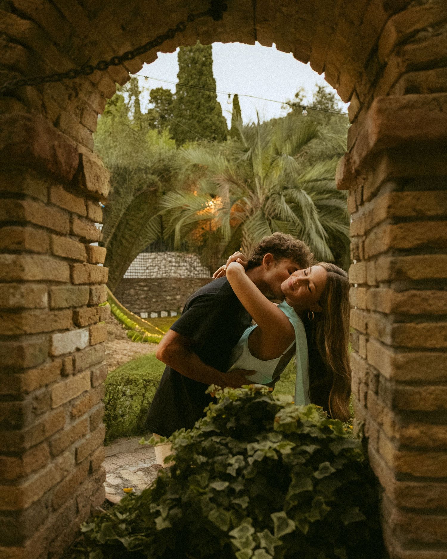 Couple shares an embrace beneath a rustic brick archway framed by ivy, palm trees, and lush greenery in the background.