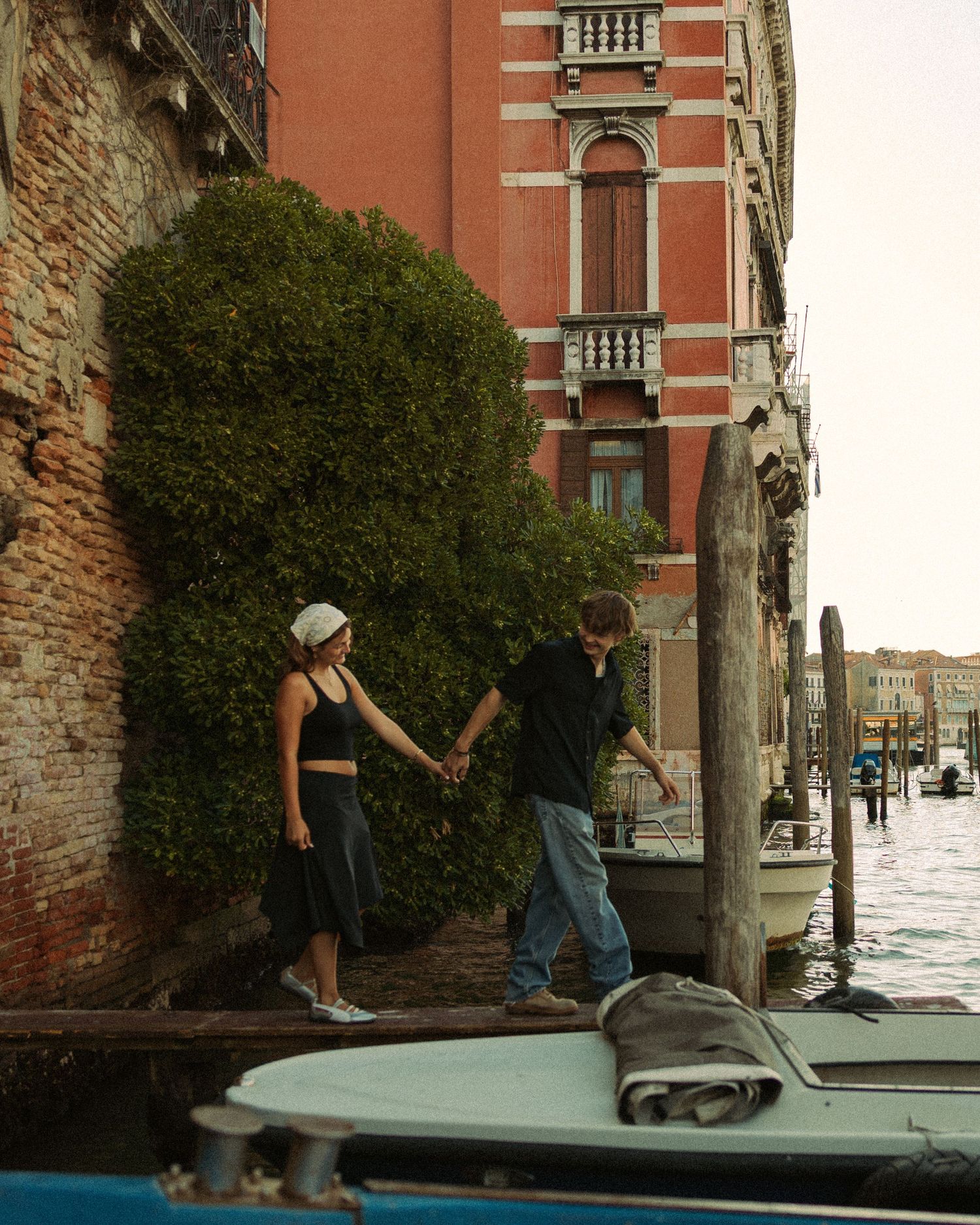 Two people hold hands while stepping from a wooden dock onto a boat beside an ivy-covered brick wall on a Venice canal.