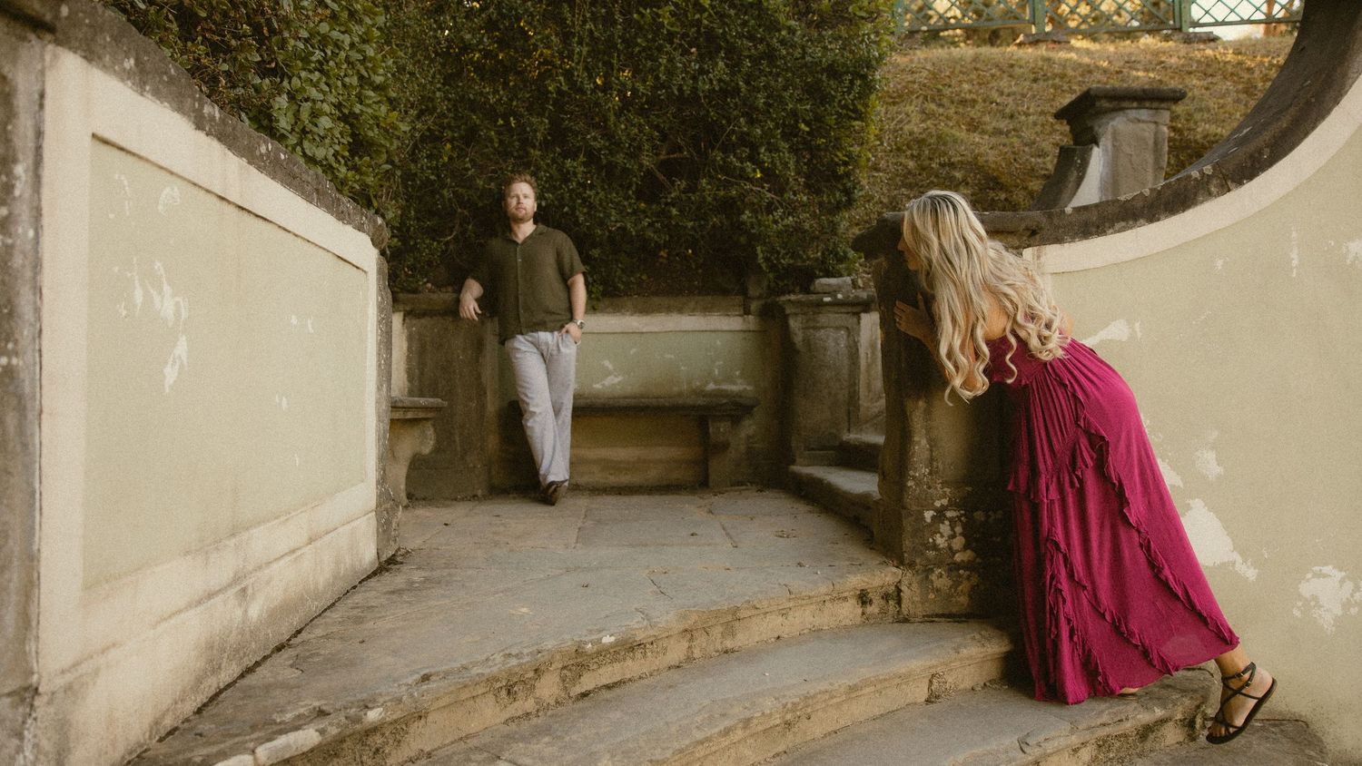 A woman in a flowing red skirt climbs stone steps toward a man waiting on a garden terrace surrounded by lush greenery.