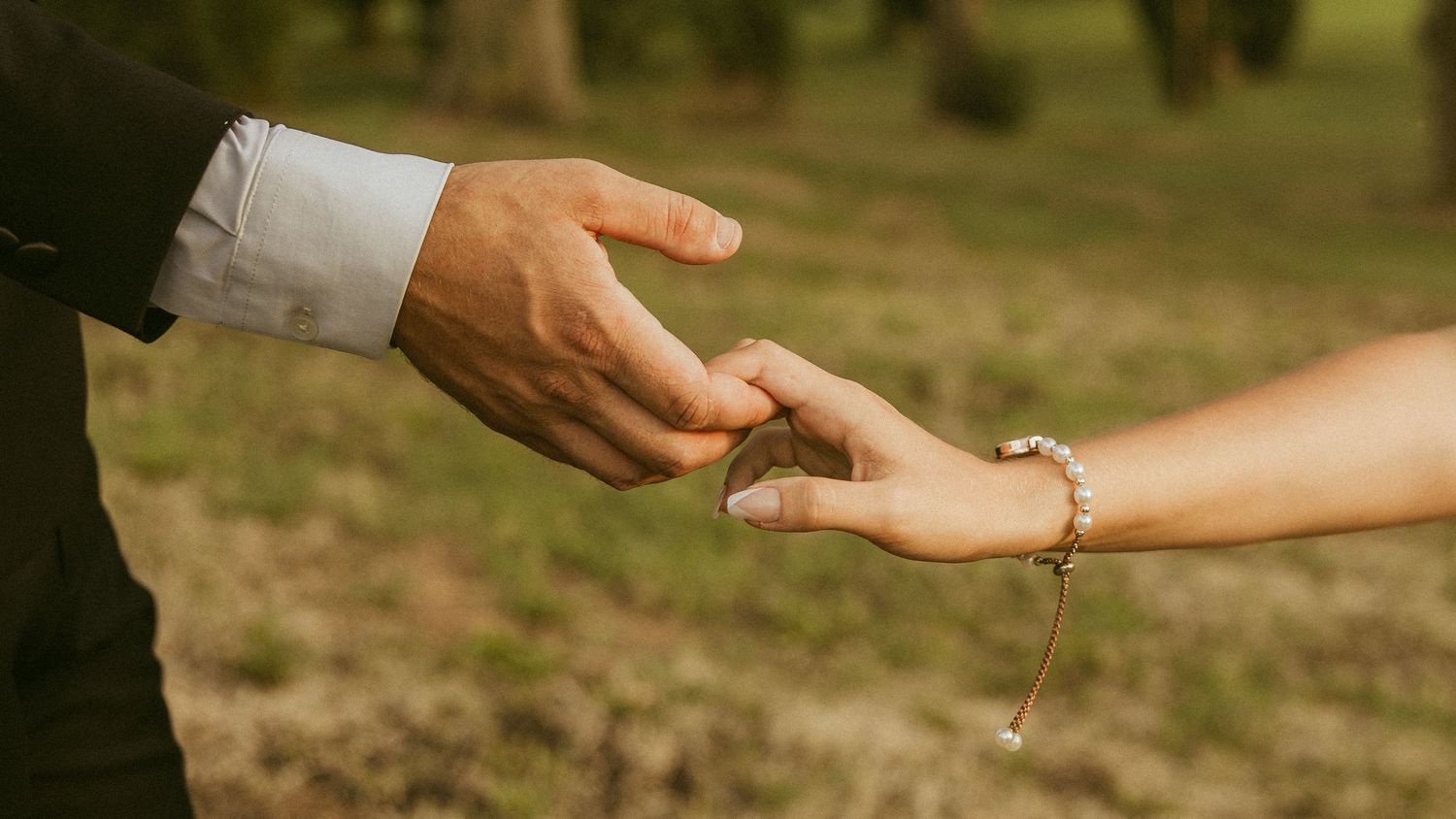 Man in a dark suit and white cuffed shirt gently holds the hand of a woman wearing a beaded bracelet outdoors on a grassy lawn.