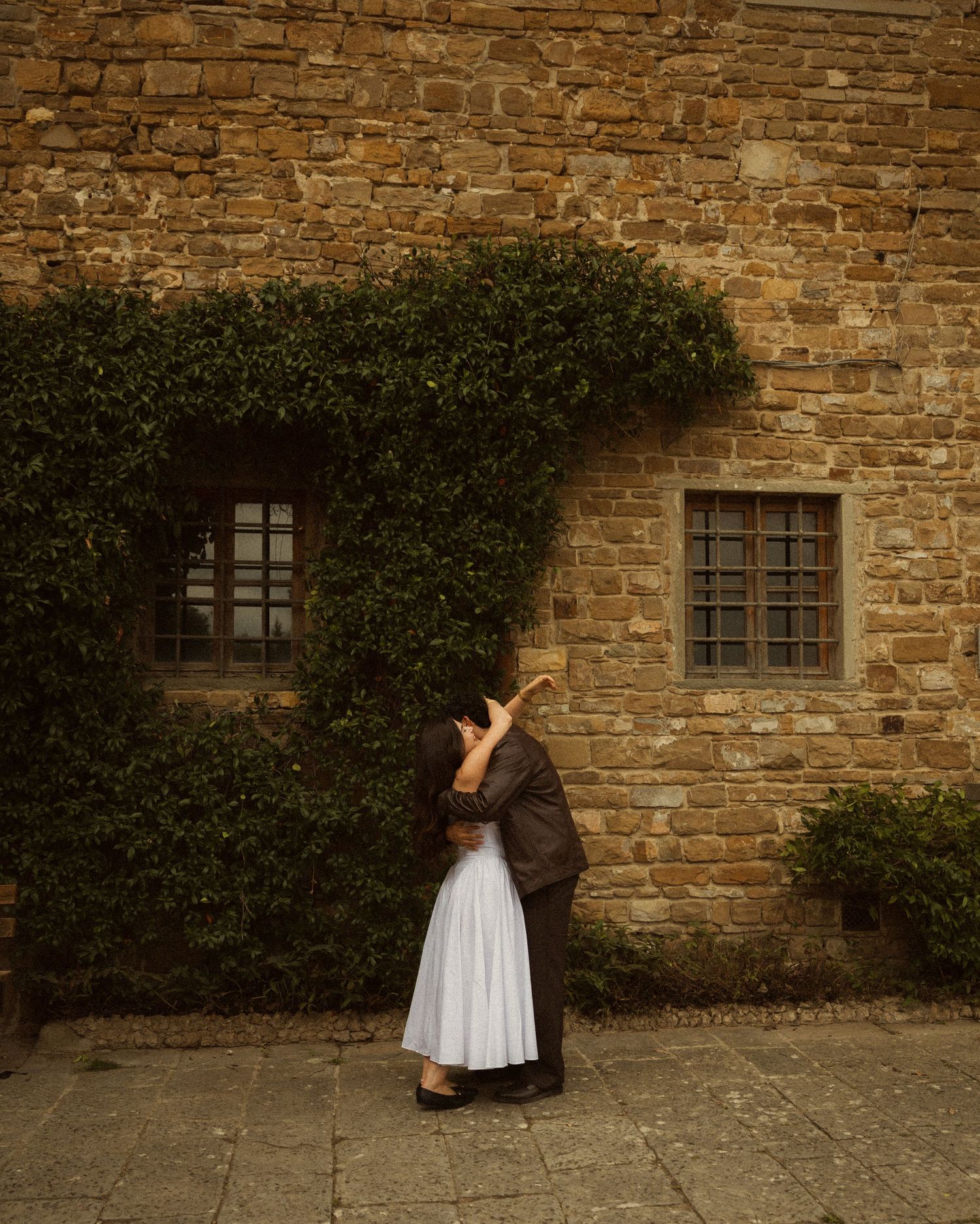Couple kissing in front of a rustic brick building with ivy-covered windows during an engagement photo session. ROMANTIC COUPLE IN FLORENCE