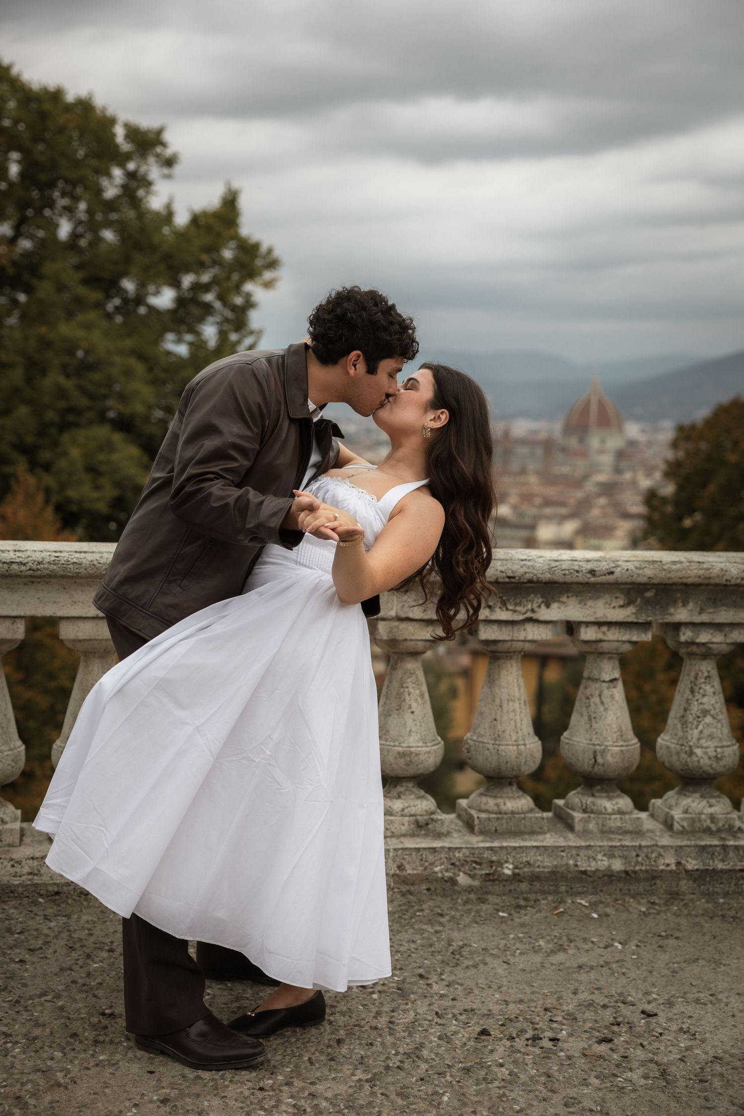 Couple in a romantic dip kiss on a stone balustrade, the bride in a white tea-length dress, with a hilltop city view and dome behind.