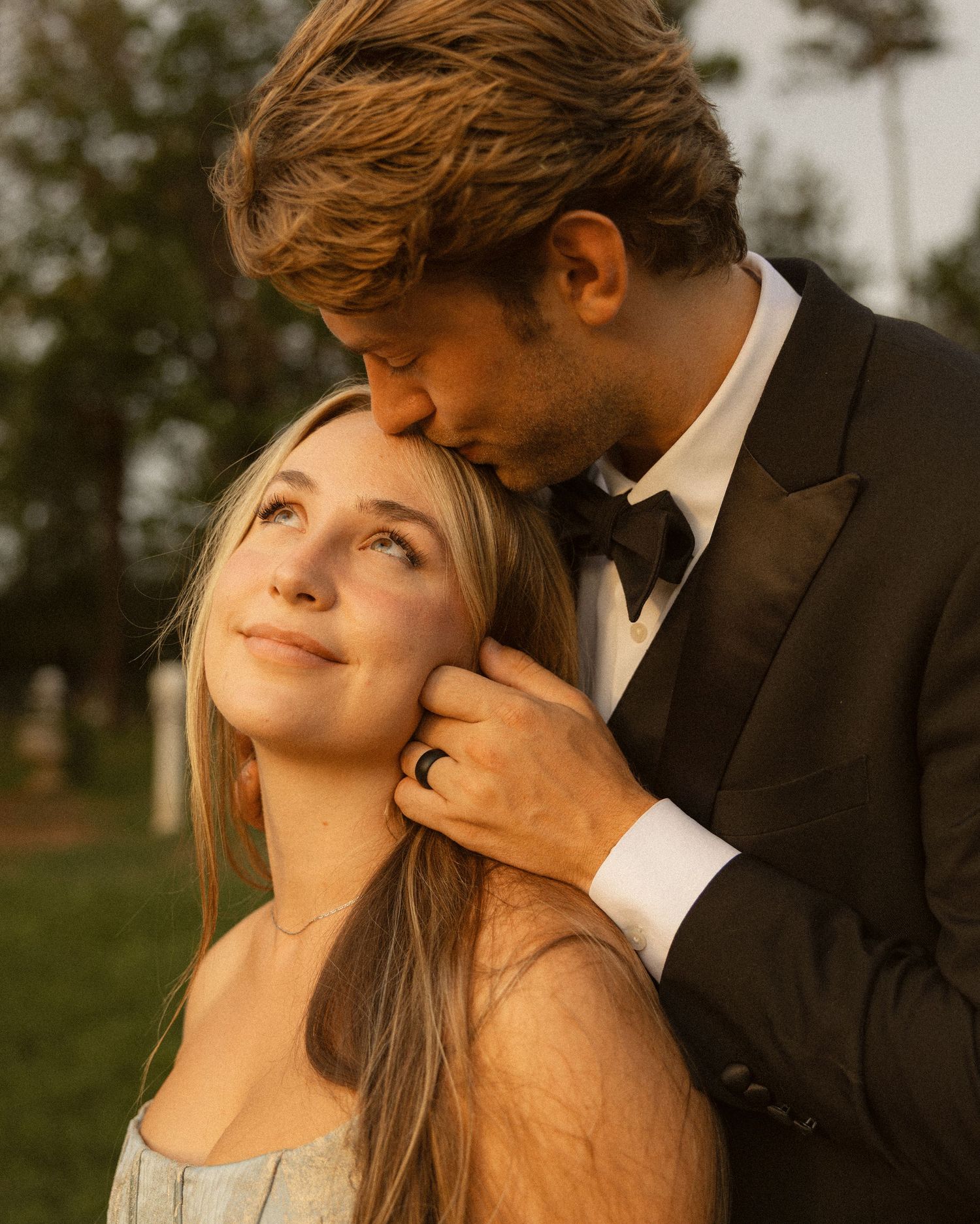 Groom in a black tuxedo and bow tie kisses the forehead of a smiling bride in a sage green dress during a romantic outdoor wedding portrait.
