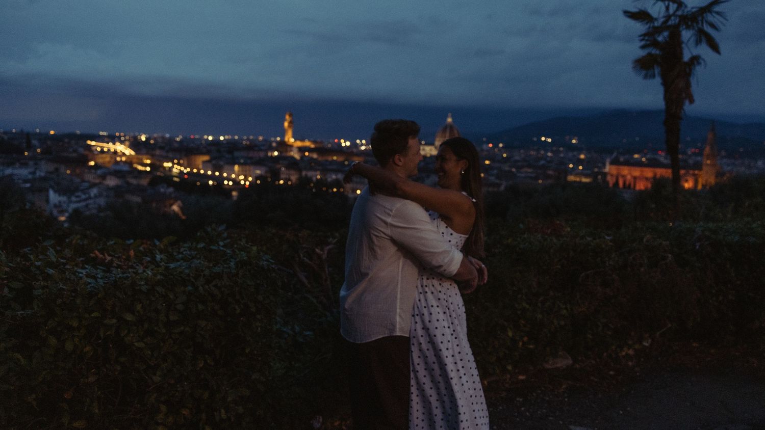 A couple embraces at a hillside overlook at dusk, with a glowing city skyline and distant domes twinkling below a deep blue evening sky.