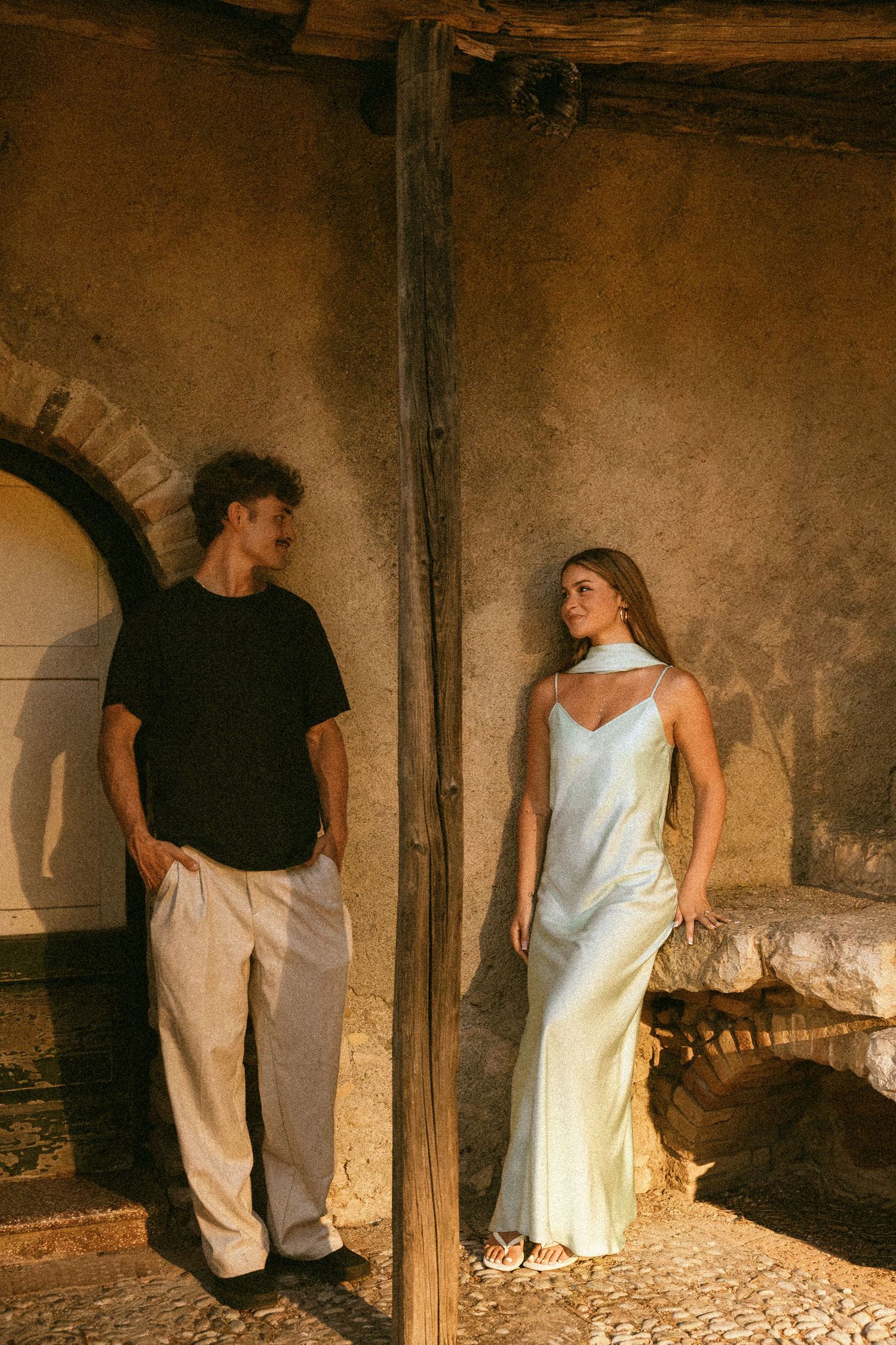 A man in a black t-shirt and linen pants leans against a wooden post beside a woman in a satin slip dress in warm golden light.