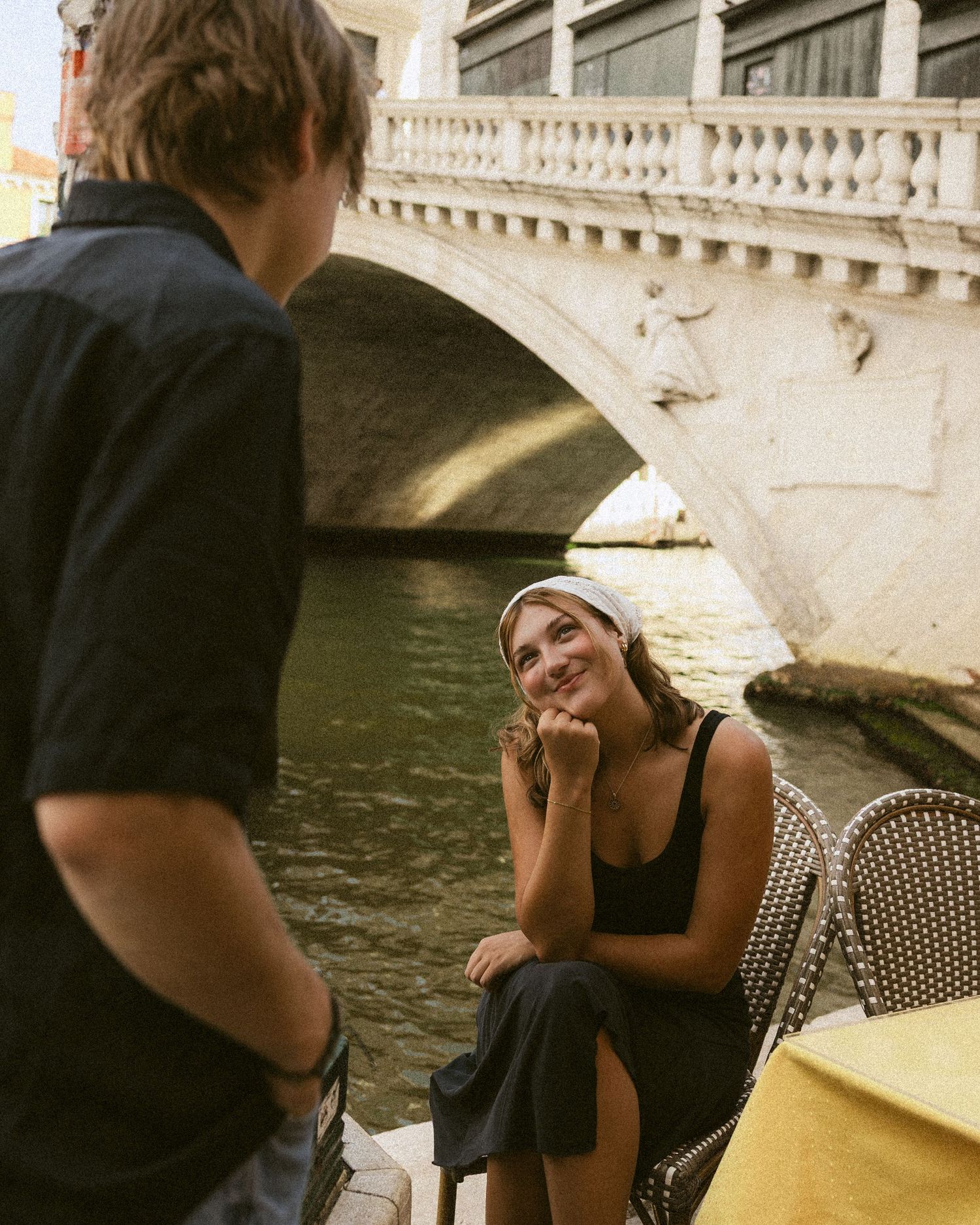 Smiling woman in a white headscarf rests her chin on her hand at a canalside ccafé table while a man talks, with a stone bridge behind.