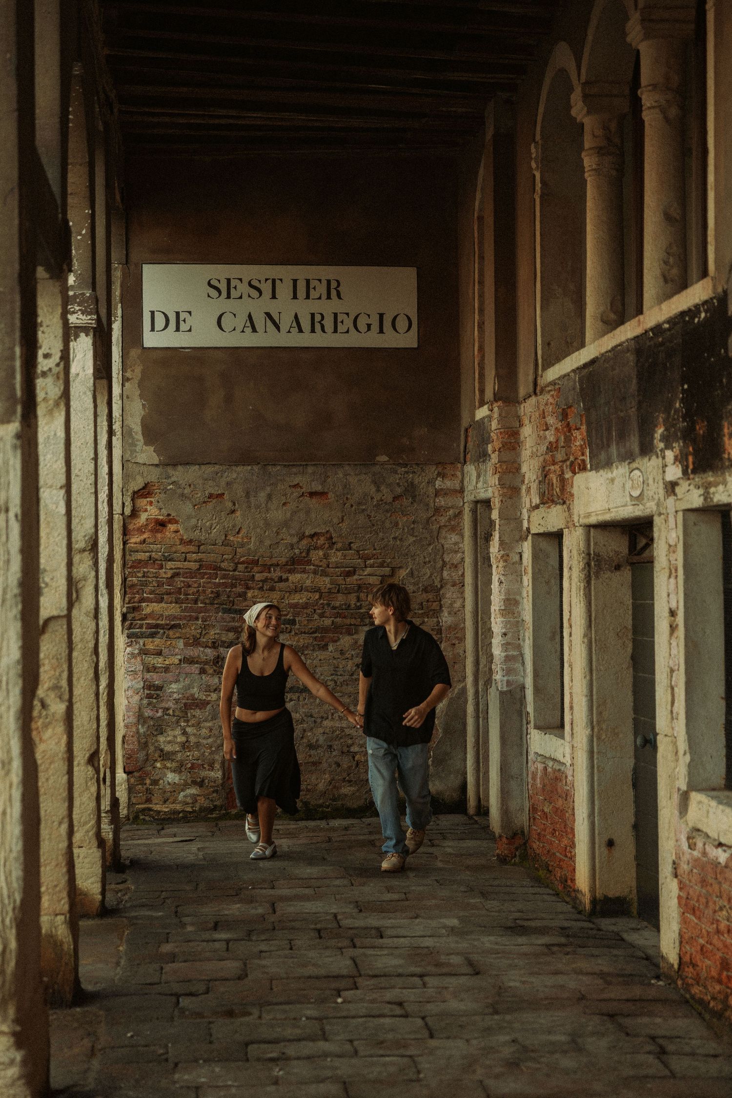 Couple walking hand in hand through a weathered brick archway in Venice beneath a Sestier de Canaregio sign.