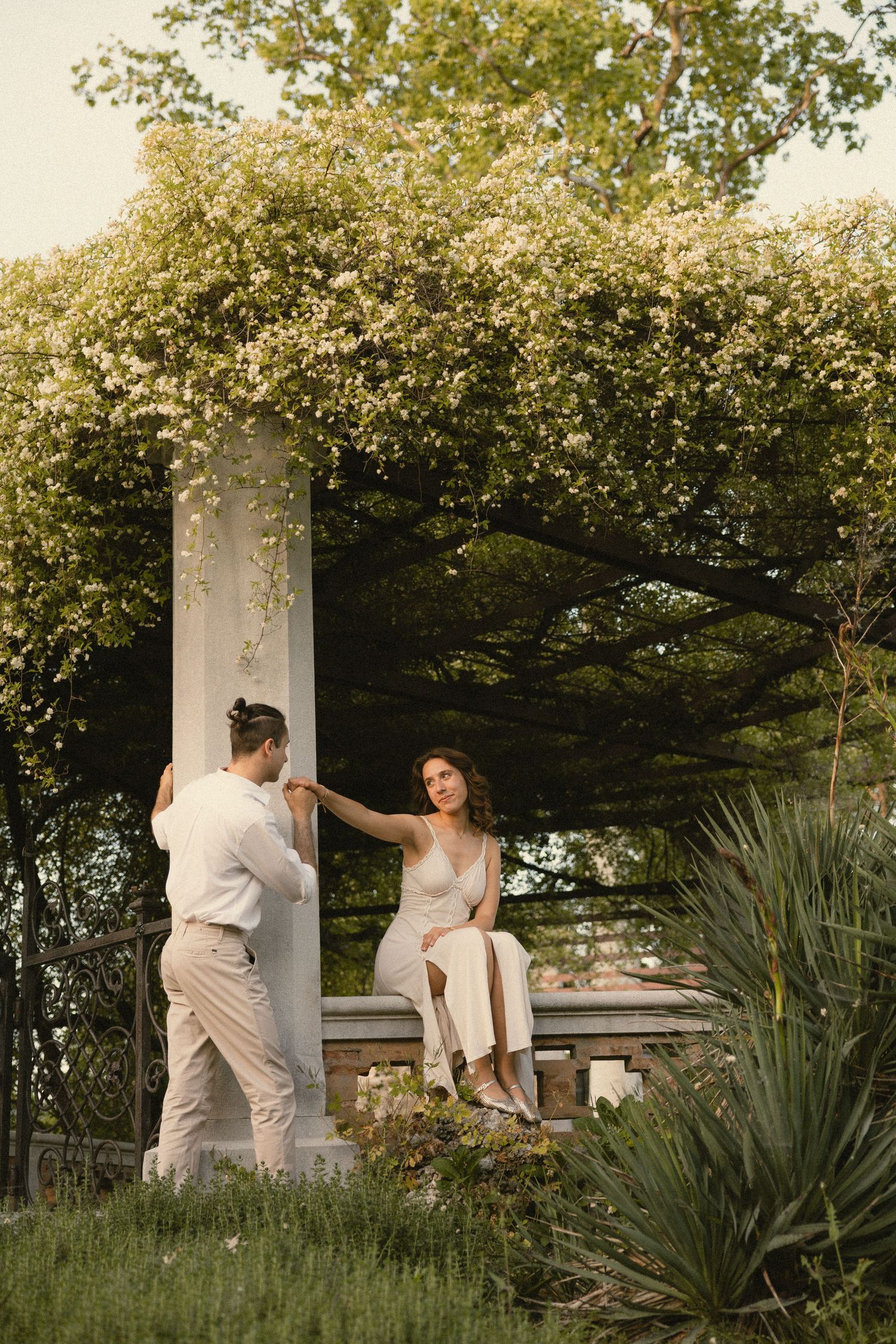 Couple in white outfits holding hands at a stone pergola draped in blooming white vines, perfect for a romantic engagement photo shoot.