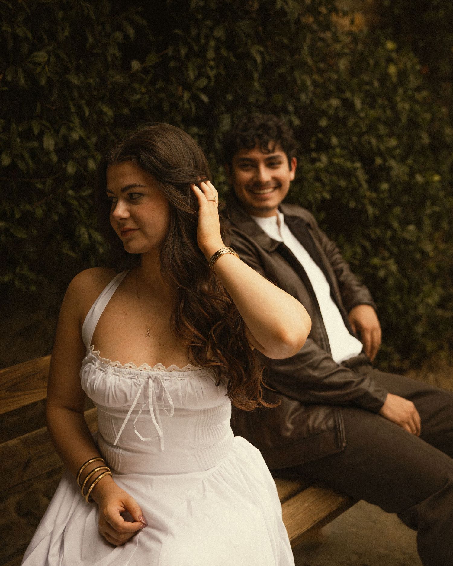 Smiling man in a leather jacket sits on a wooden bench behind a woman in a white sundress touching her hair. ROMANTIC COUPLE IN FLORENCE