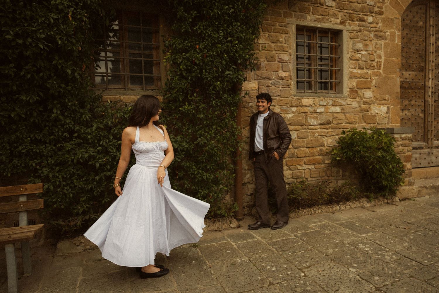 Woman in a flowing white dress twirls on a stone courtyard while a man in a dark jacket leans against a rustic ivy-covered brick wall.