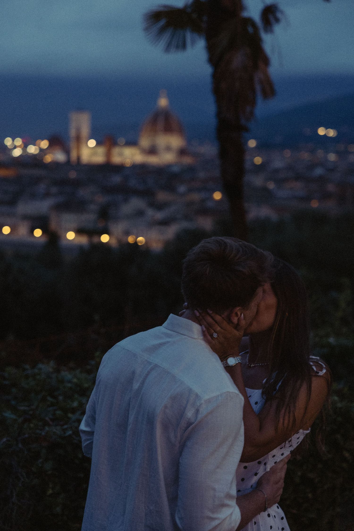 Couple kissing at dusk on a hillside overlooking Florence, with the illuminated Duomo and city lights glowing beneath a palm tree.