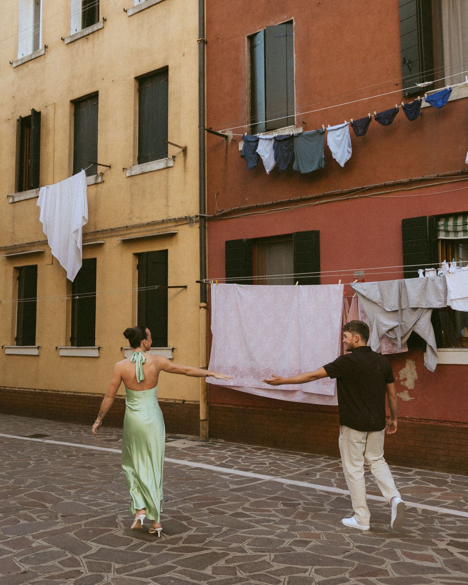 Couple in a green satin dress and black shirt reach for each other on a cobblestone street beneath laundry lines in Venice.