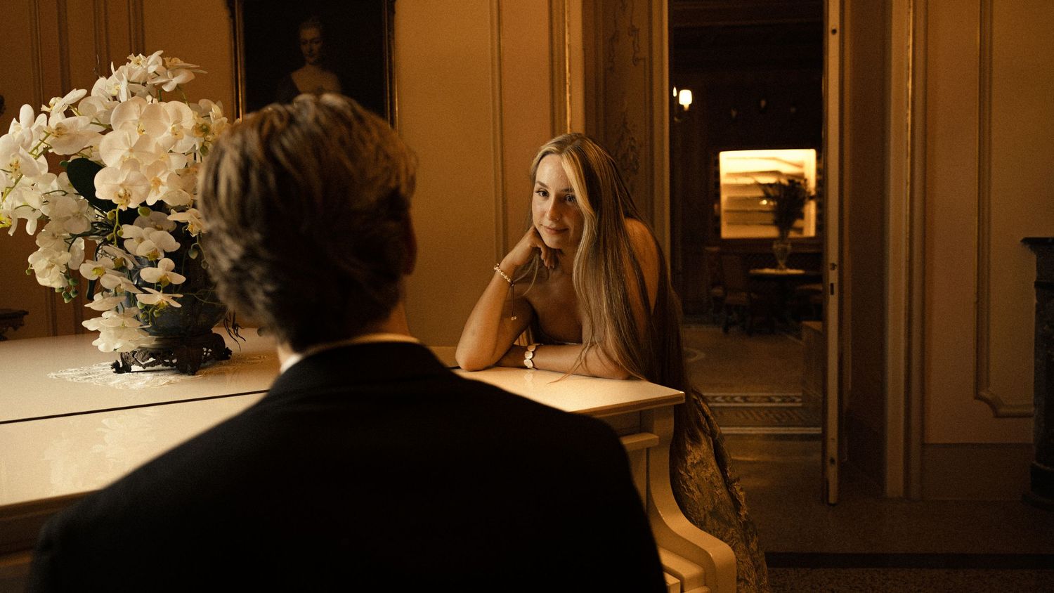 Long-haired woman in a white dress smiles from a table in an elegant room with white flowers while a man sits facing her in the foreground.