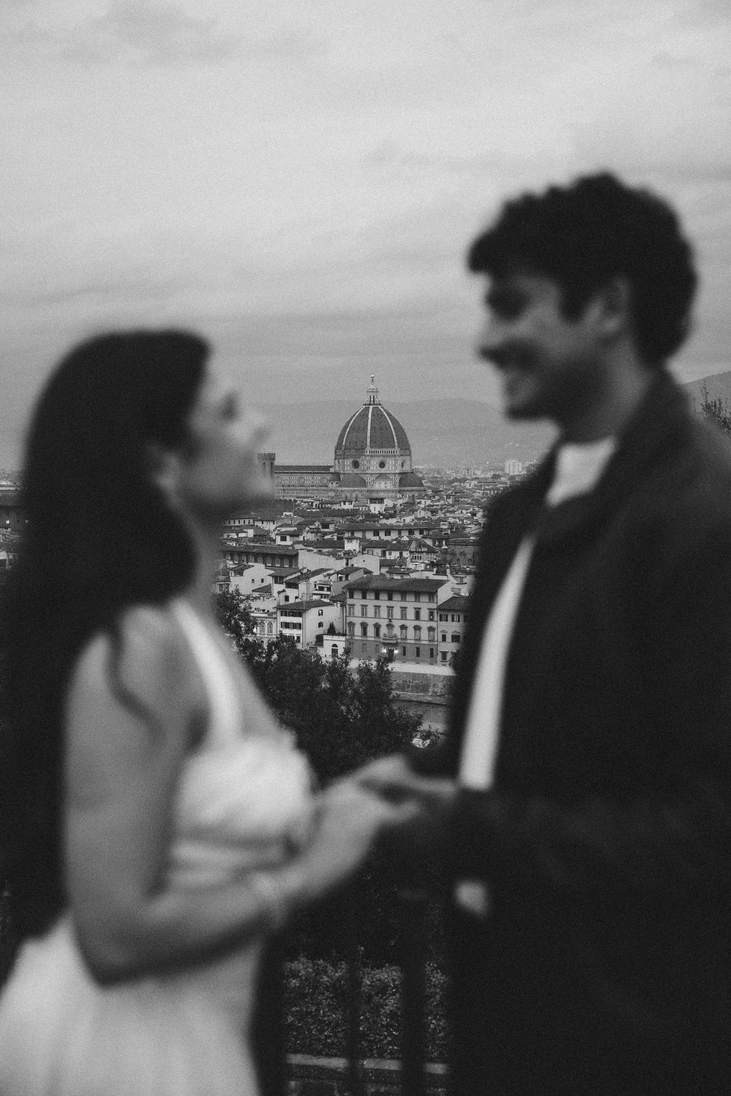 Black and white photo of a couple holding hands on a overlook with the Florence Duomo and city rooftops blurred in the background.