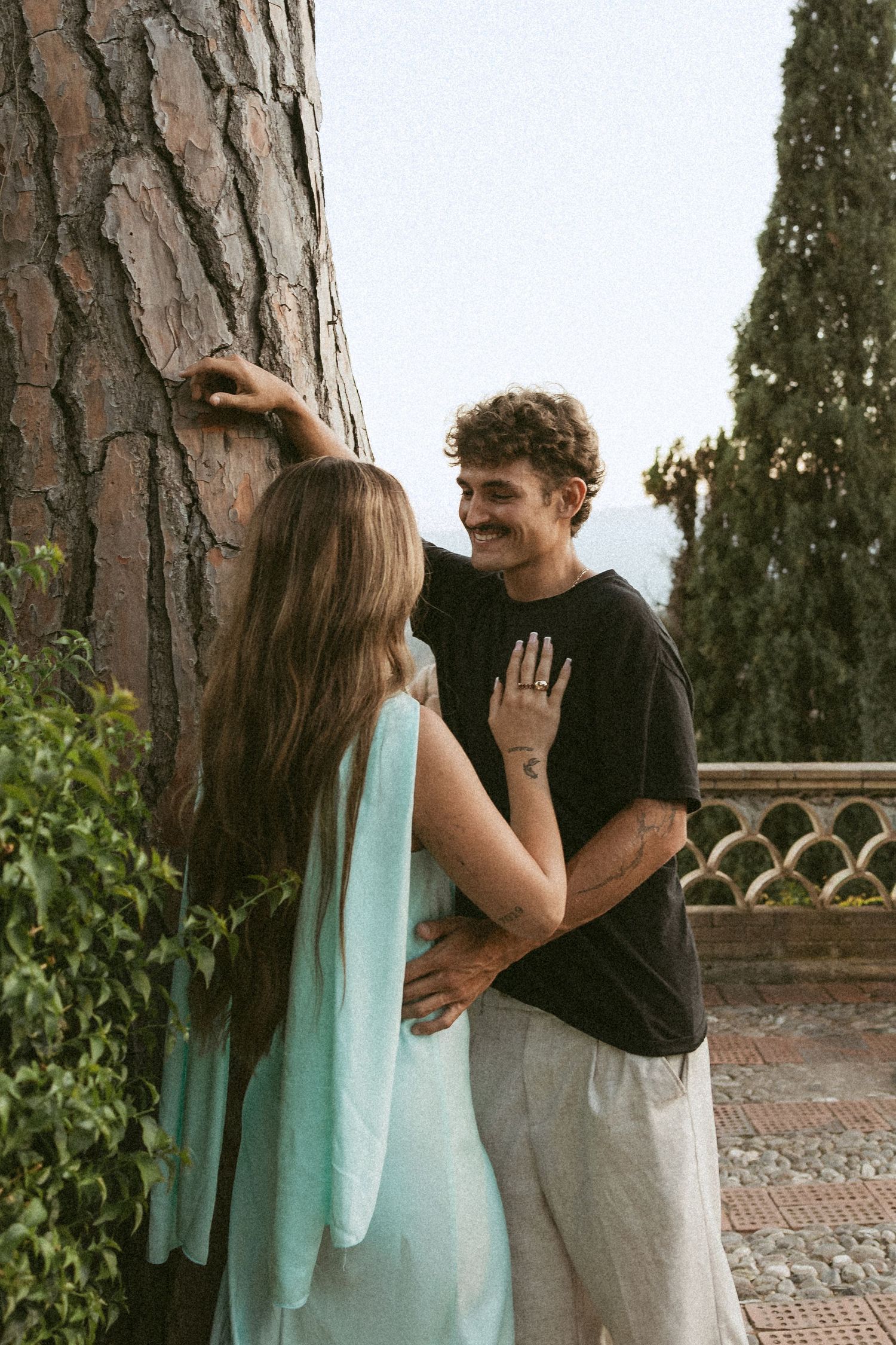 Smiling couple embraces beside a large pine tree on a terrace, the woman in a flowing mint green dress and the man in a black tee.