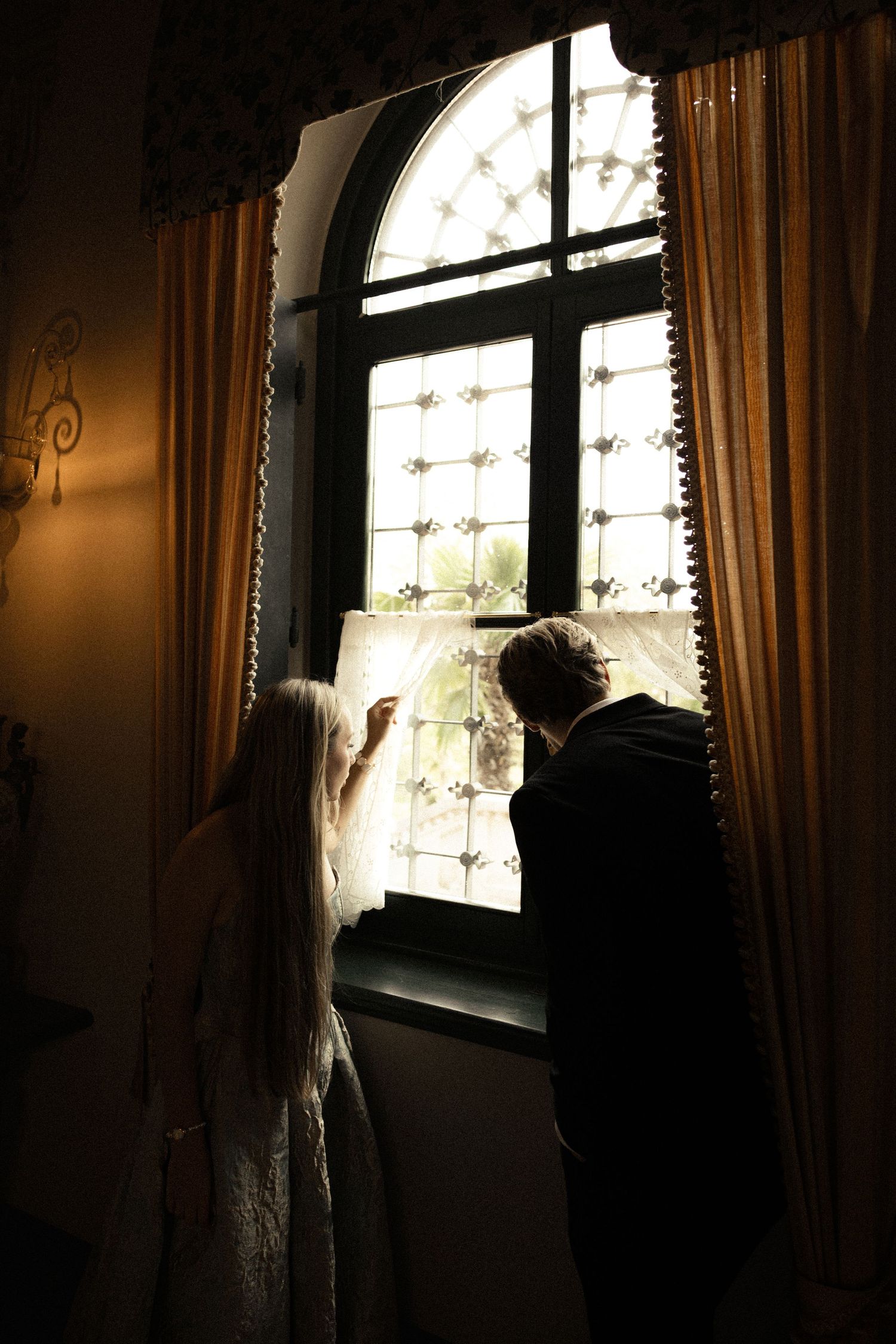 Silhouetted bride in a lace gown and groom in a dark suit gaze out an arched window framed by golden curtains.