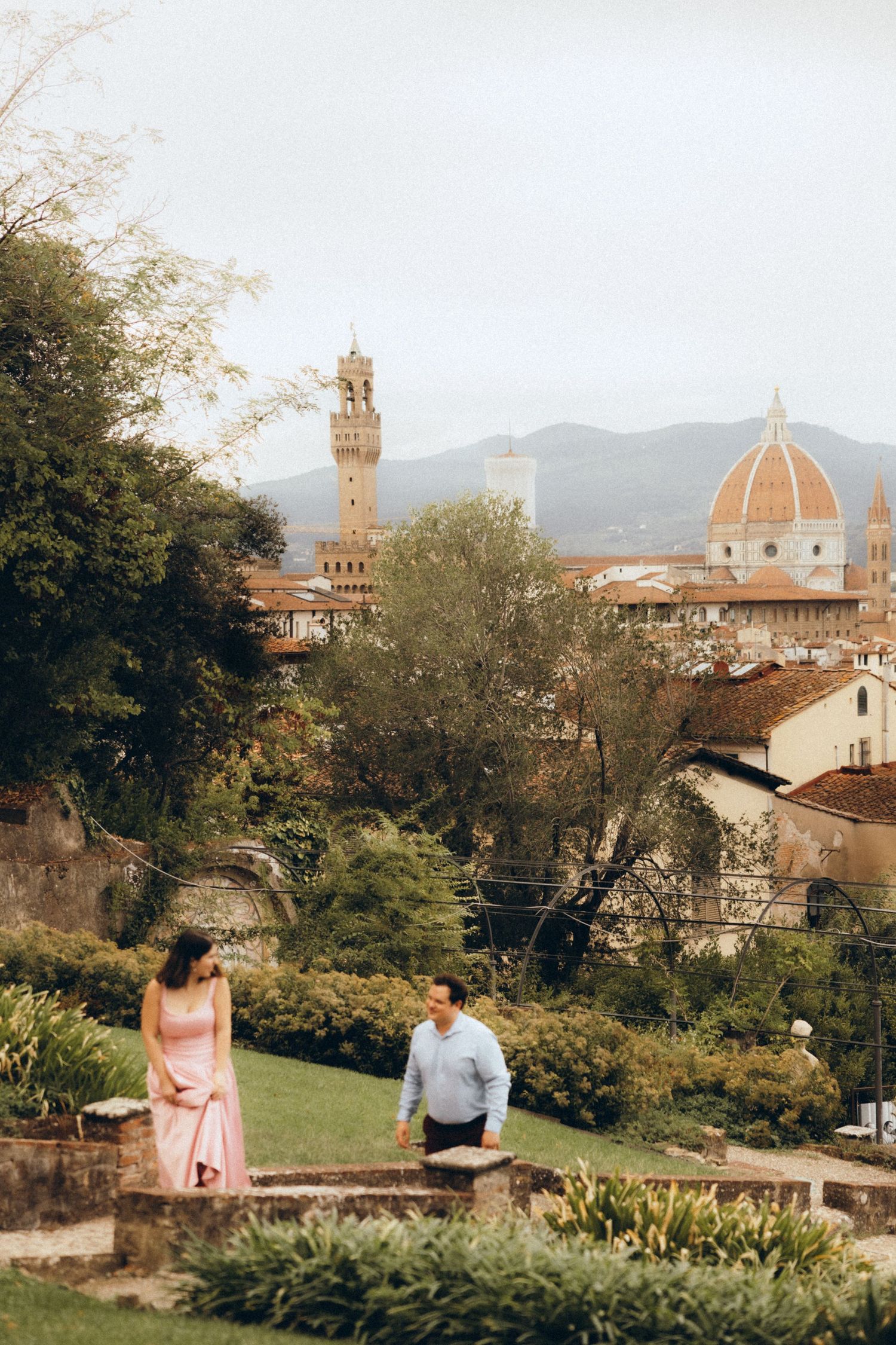 Couple standing in a garden overlooking Florence, with the Duomo dome and Palazzo Vecchio tower rising above terracotta rooftops.