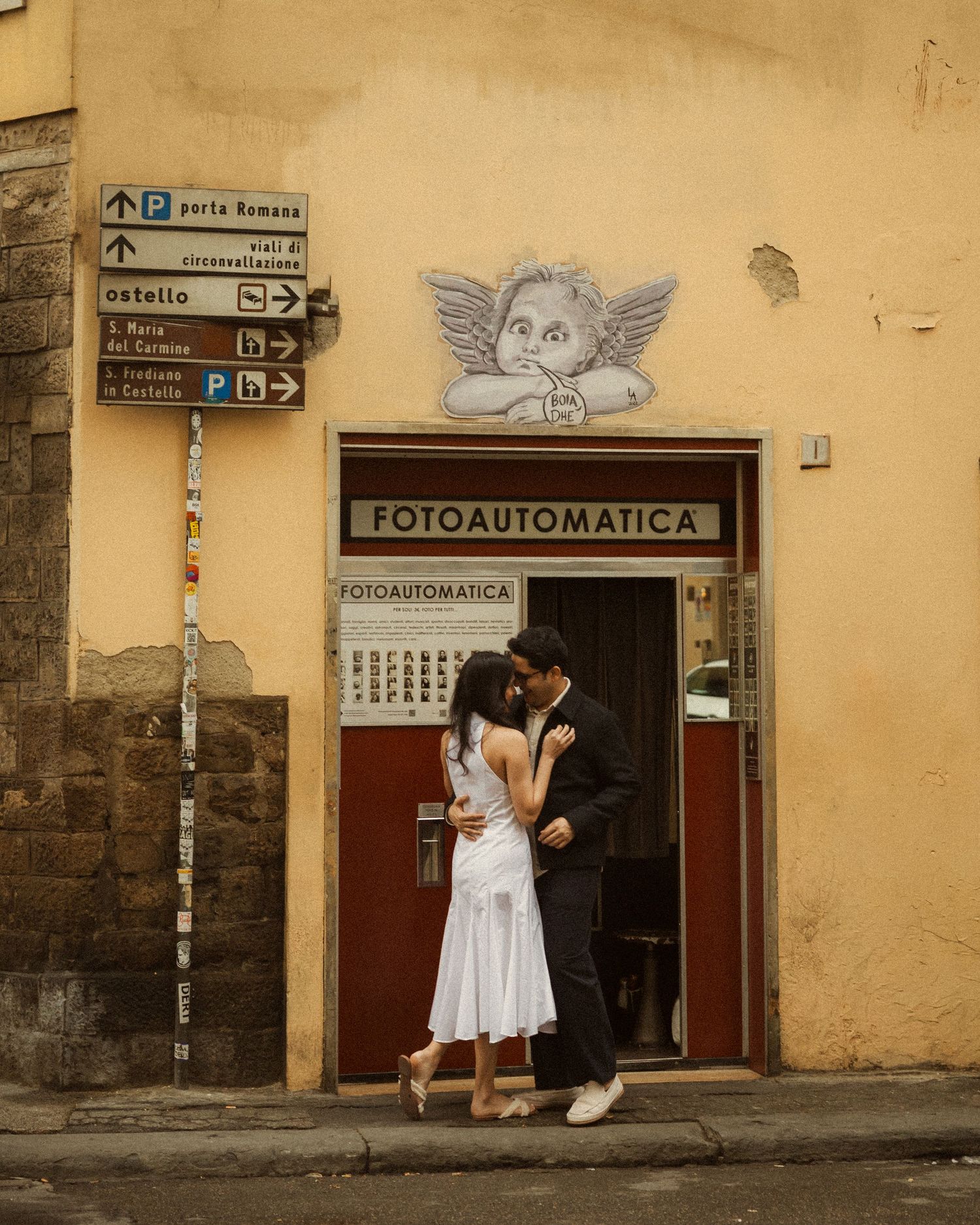 Couple embracing in front of a vintage Fotoautomatica photo booth in Florence with street signs and an angel relief above. ROMANTIC FLORENCE
