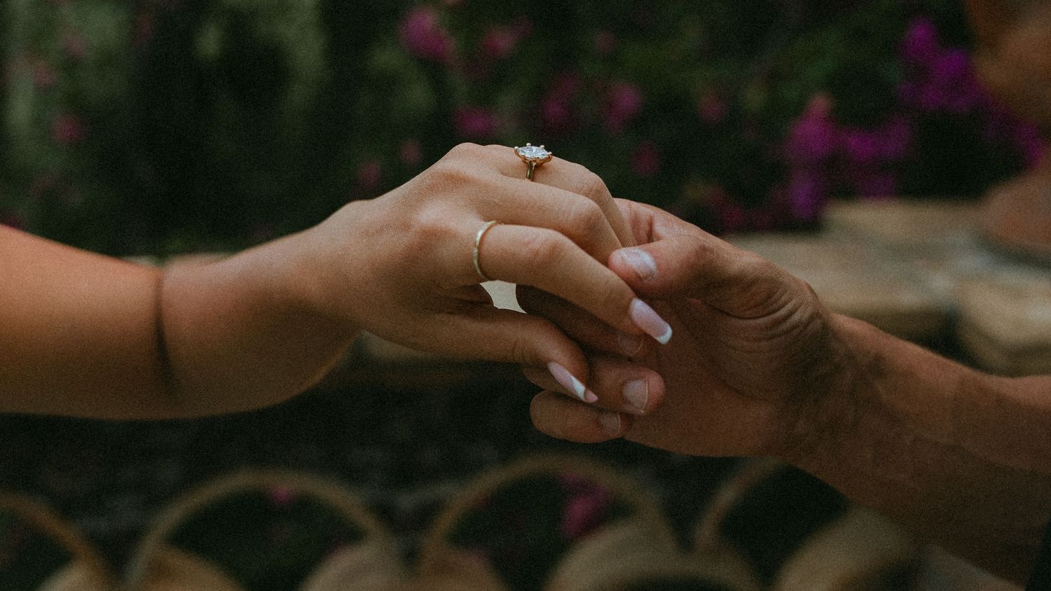 Two hands gently clasped together, one wearing a diamond engagement ring, against a blurred garden background with purple flowers.