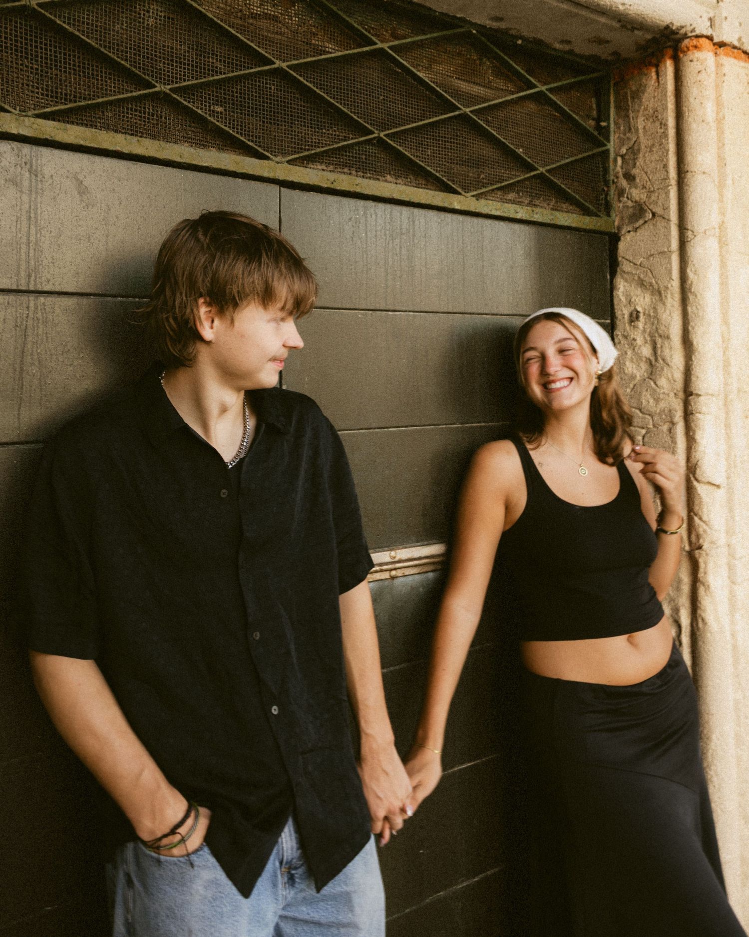 Young couple holding hands and laughing together, leaning against a rustic wooden wall in warm, golden light.